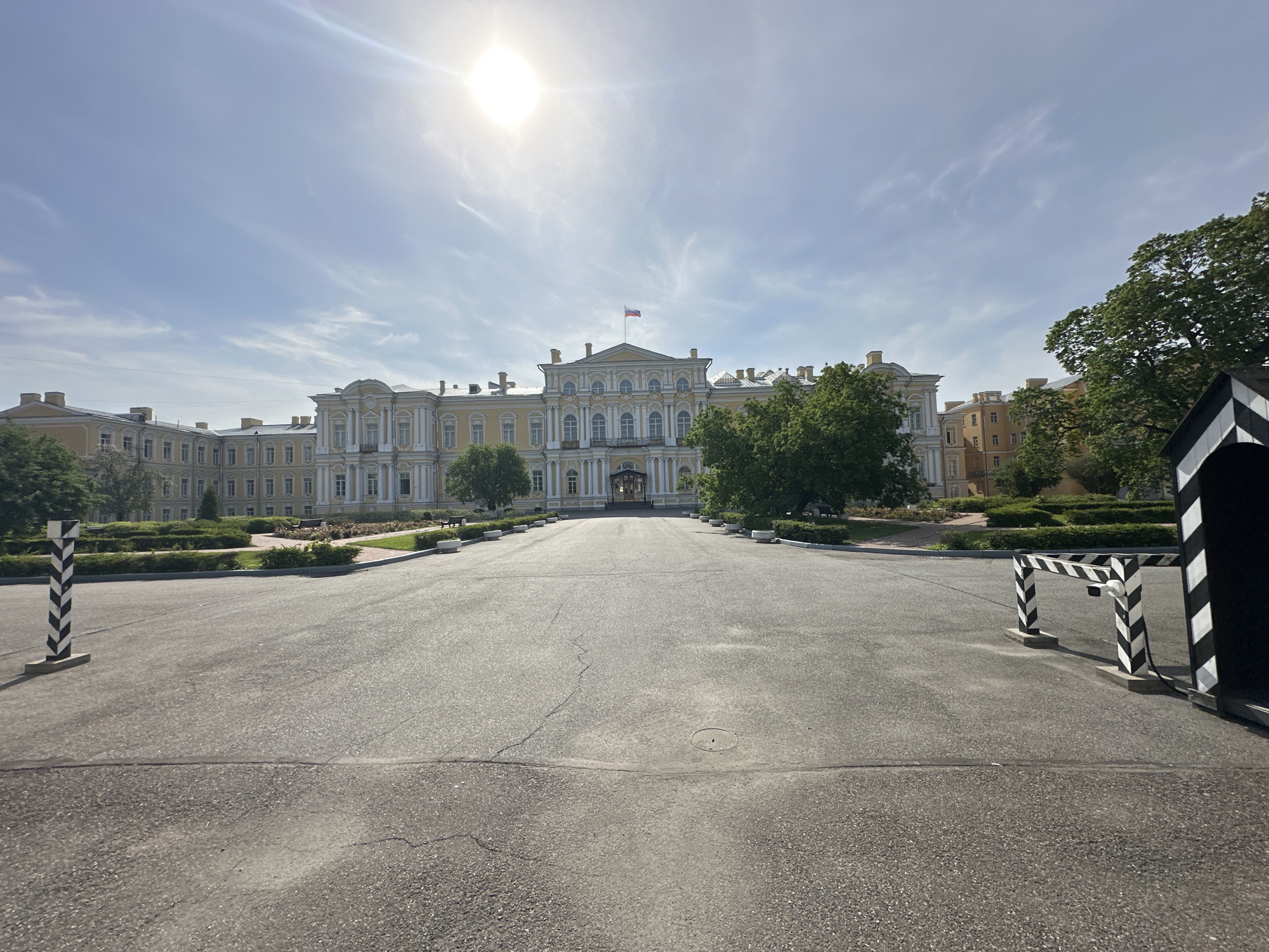 This image depicts the Konstantinovsky Palace in Strelna, Saint Petersburg, Russia, as viewed from a paved approach. The sky is partly cloudy with a visible sun. The palace is a light-colored building with ornate detailing and a Russian flag on its roof. Manicured hedges and trees flank the roadway leading to the palace entrance. In the foreground are elements of a guard post or gate, with black and white chevron patterns. The road surface is asphalt, showing some cracks and signs of wear. There is no visible human presence in the immediate scene.
