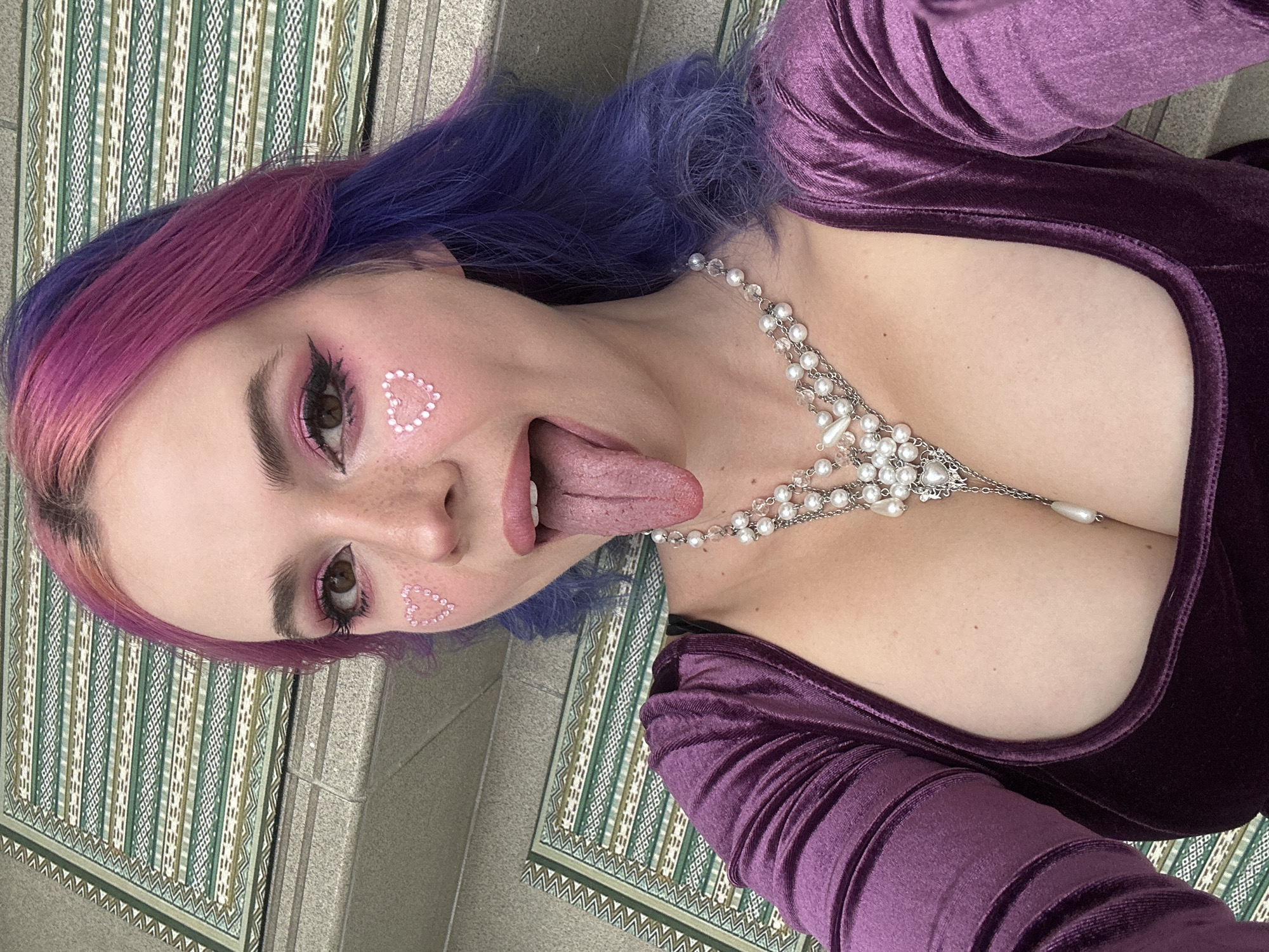 A woman with pink and blue hair is lying on a patterned surface, possibly a rug or mat, against a grey background that may be concrete. She is wearing a purple velvet dress and a pearl necklace that draws attention to her chest. The woman has heart-shaped makeup on her face and is sticking her tongue out. The photo appears to be taken at a close angle, focusing on her upper body and face. There are no apparent indicators within the image to suggest the City of Zagreb, Croatia.
