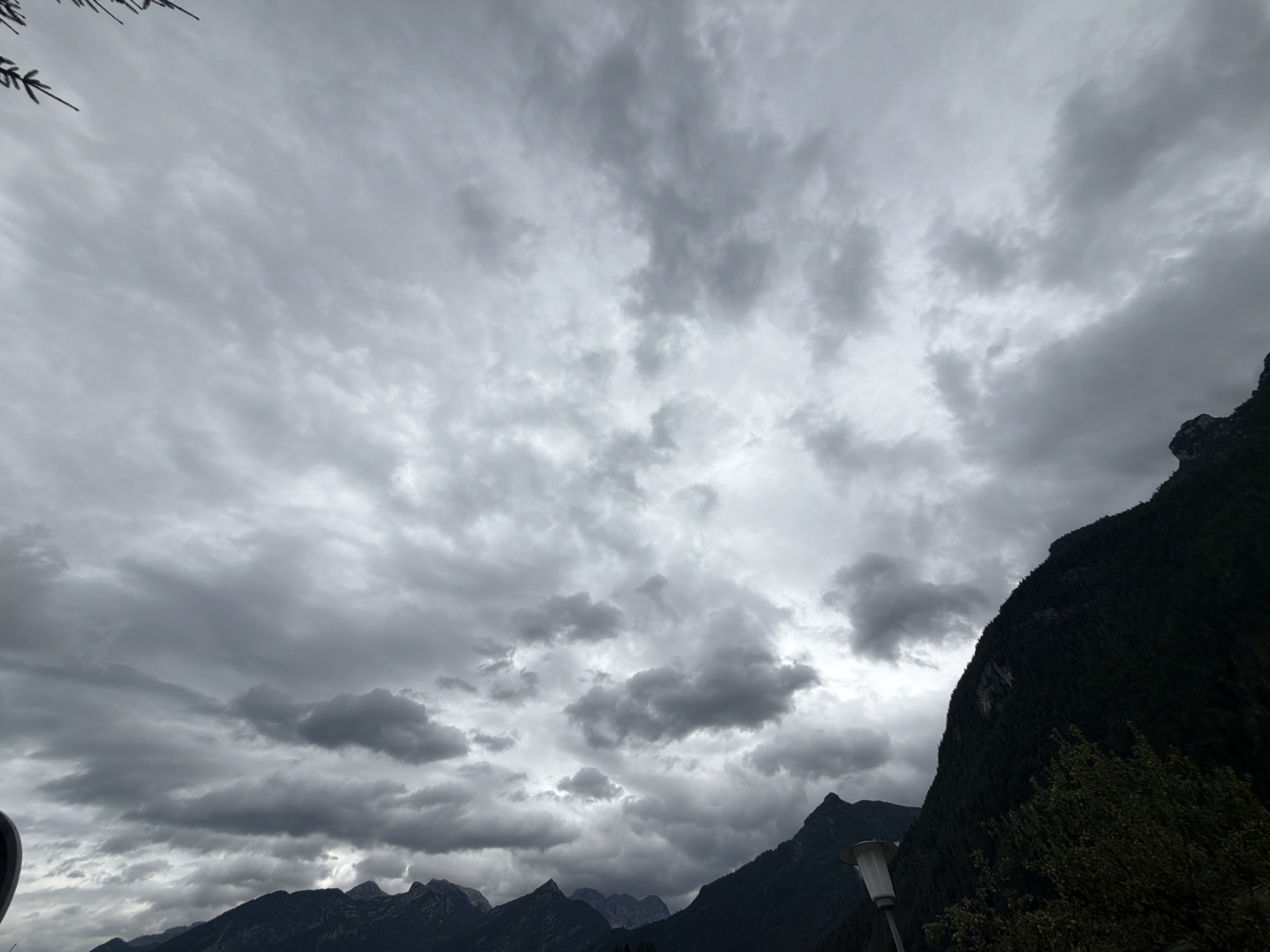 The image depicts a dramatic, cloudy sky over a mountainous landscape. The sky is filled with dark, ominous clouds, suggesting overcast or stormy weather. The clouds are textured and varied, with some appearing heavy and gray, while others are lighter and more wispy.
In the foreground, the dark silhouettes of mountain ranges are visible, with the slopes covered in dense vegetation, likely trees. The peaks of the mountains are sharp and rugged. To the right, a steep, dark mountainside dominates the frame, also covered in foliage. A modern-looking streetlamp with a white top is visible in the lower right portion of the image, indicating a man-made element within the natural setting. A few dark branches or twigs are also visible in the upper left corner, possibly from a tree or foliage in the immediate foreground.
The overall mood is somber and moody, amplified by the dark clouds and the imposing mountains. The time of day is unclear due to the heavy cloud cover, but the lighting suggests it is either early morning or late afternoon, or a day with perpetually dim light. The location is identified as Torren, Austria, which is consistent with a mountainous European setting. There is no visible text in the image.