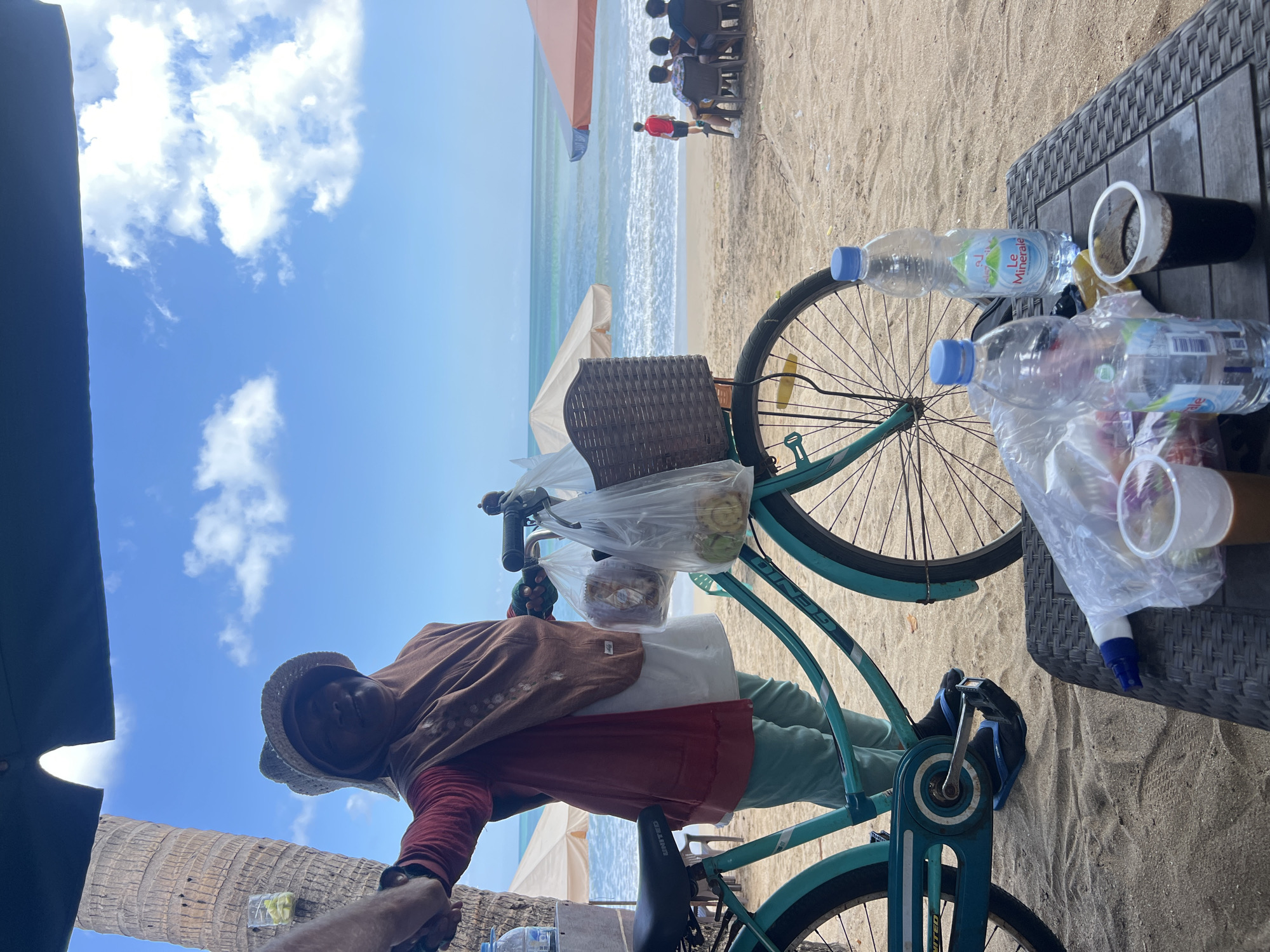 A woman selling goods on a beach in Kuta, Indonesia, is captured in a vertical, eye-level shot. She stands behind a turquoise bicycle with a basket laden with plastic-wrapped items. The woman wears a rust-colored top, a brown headscarf, and light green pants, with a straw hat atop her head. She holds a hand out, possibly to a customer.
The beach is sandy, with the ocean and a blue sky dotted with white clouds in the background. Several beach umbrellas, some occupied by people, are visible along the shoreline. To the right of the frame, a woven table holds two plastic cups, one with a dark beverage, and two plastic water bottles. The scene conveys a typical beach vendor's setup in a popular tourist destination.