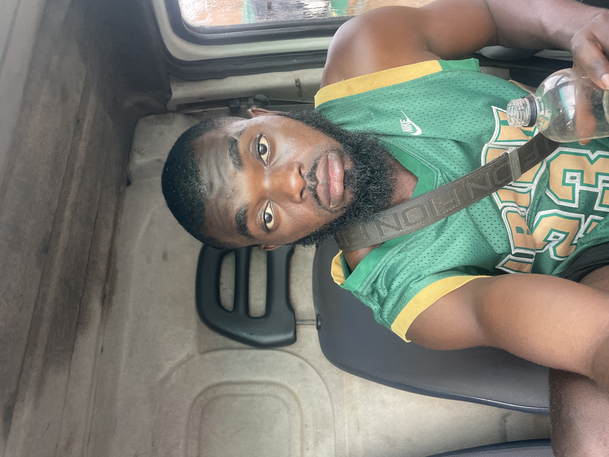 A man with dark skin, a beard, and short dark hair sits in the driver's seat of a vehicle. He is wearing a green basketball jersey with yellow trim and a black strap across his chest. He is holding a clear plastic bottle in his right hand. The interior of the vehicle is visible behind him, including a headrest and part of the ceiling. The image appears to have been taken inside the vehicle, possibly in the city of Jos, Nigeria, given the context.