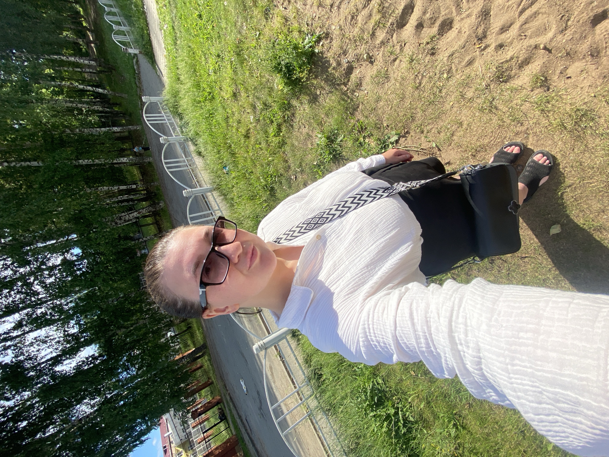 The image shows a young woman taking a selfie outdoors in Chaikovsky, Russia. She is wearing a white, crinkled, long-sleeved blouse, black pants, black sandals, and sunglasses. A black bag with a patterned strap is slung across her body. The woman is looking at the camera with a neutral expression.
The setting appears to be a park or a grassy area next to a paved path. There are trees with white bark, likely birches, visible in the background, suggesting a summer or spring day. The ground is covered with dry grass and dirt, indicating a sunny and warm day. A white, decorative metal fence runs along the path. In the far background, buildings and a black fence are visible. The sun is shining brightly, casting shadows and creating a warm atmosphere. The time of day appears to be daytime, likely afternoon, due to the angle of the sun and the strong shadows.