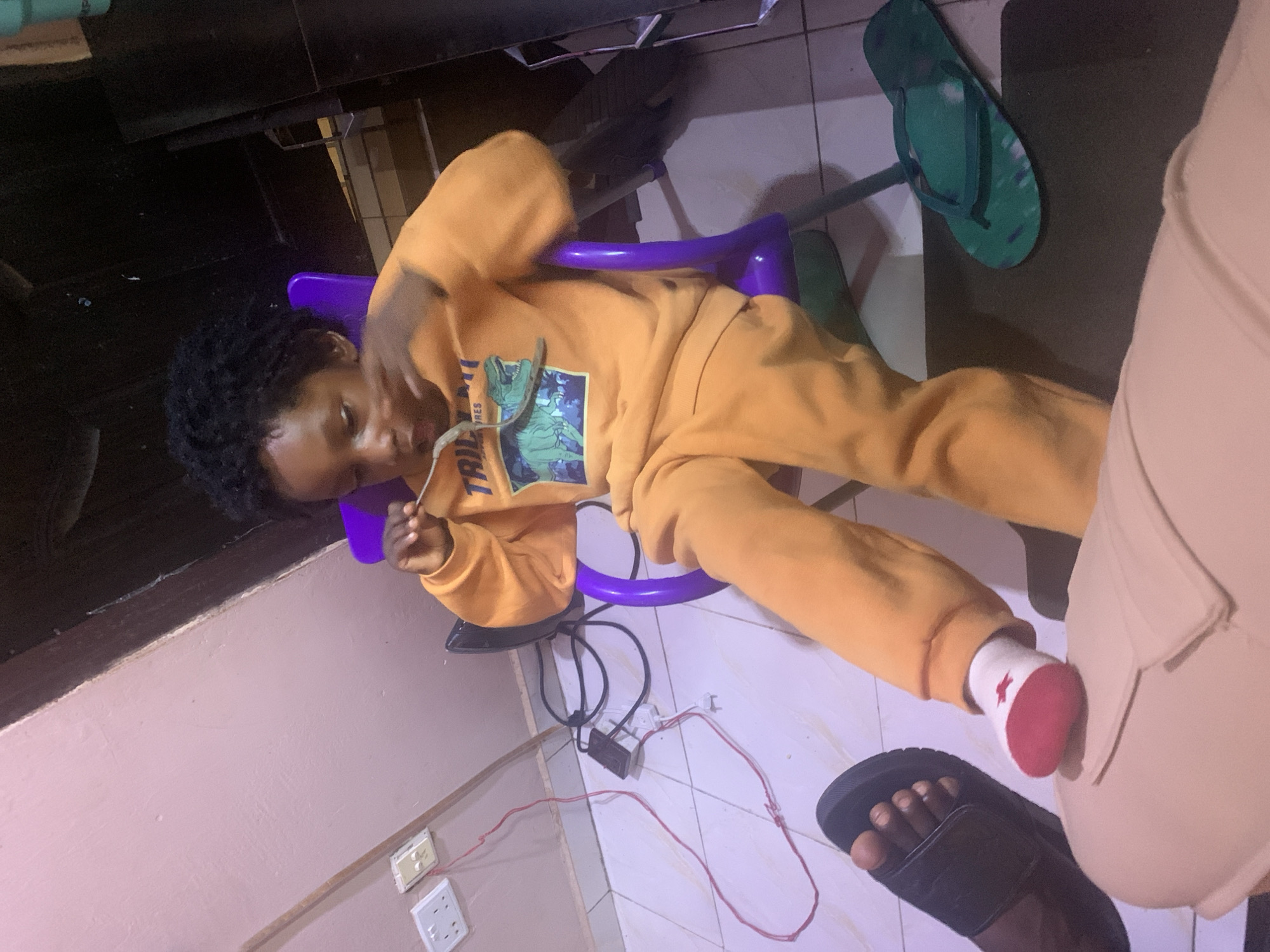 A child wearing an orange sweatsuit with a dinosaur design is reclining in a small, purple plastic chair. The child is positioned in a room with light purple walls and tiled floors. On the floor, there are electrical outlets, wires, and plugs. A black sandal and a green flip-flop are also on the floor near the child. There is a person in the bottom right corner of the photo and the setting appears to be indoors. While the precise location in Rafin Bauna, Nigeria, cannot be definitively determined without additional context, the image suggests a domestic interior.
