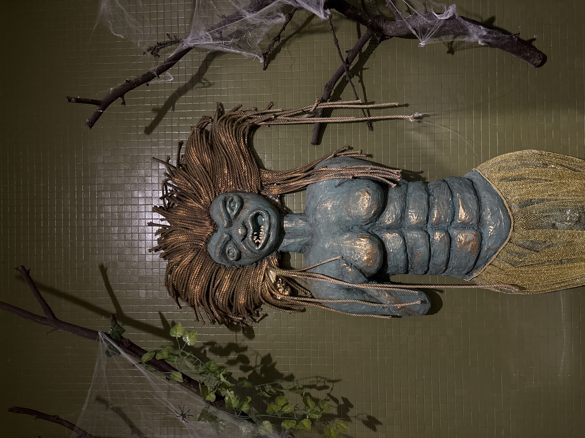The image depicts a wall-mounted Halloween decoration of a grotesque, possibly undead, mermaid figure. The figure has grey-blue skin with golden highlights, a fierce expression with bared teeth, and hair made of golden-brown rope. The tail is constructed of gold-colored netting. The decoration is mounted on a green tiled wall. Artificial spiderwebs and bare branches are arranged around the figure, completing the spooky aesthetic. There is a plant (likely artificial) with green leaves hanging down on the bottom left. The location information requested is not contained within the image.
