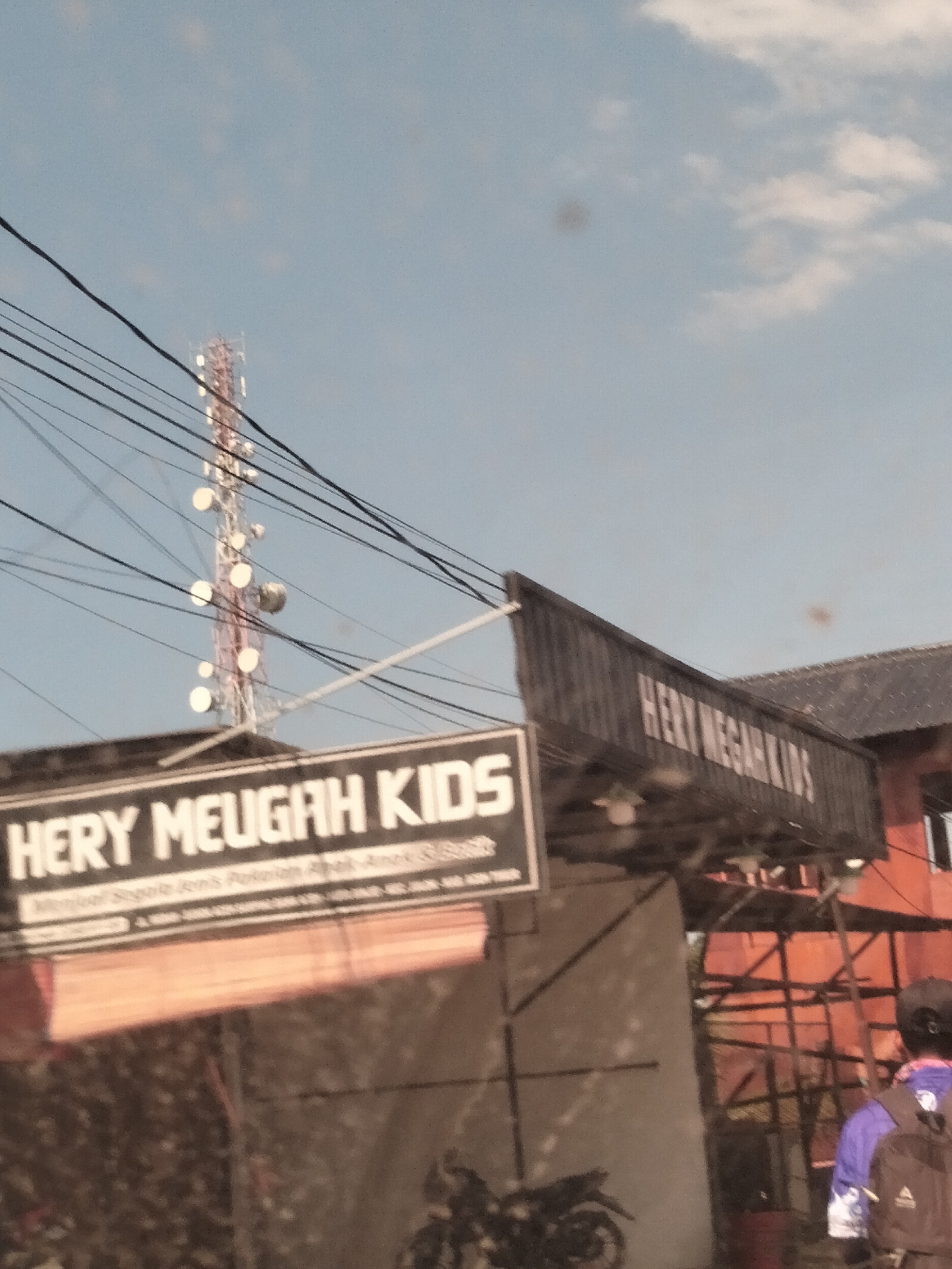 The image shows an outdoor scene in Meudangara, Indonesia. A cellular tower stands tall against a blue sky with a few clouds. There are utility wires in the foreground. Two signs are visible, both reading "HERY MEUGAH KIDS" with additional text below, possibly describing the business. A person wearing a blue shirt, a backpack, and a helmet or hat is partially visible on the right side of the image. A motorcycle is parked near the signs. Some red structural elements appear to be part of the building.