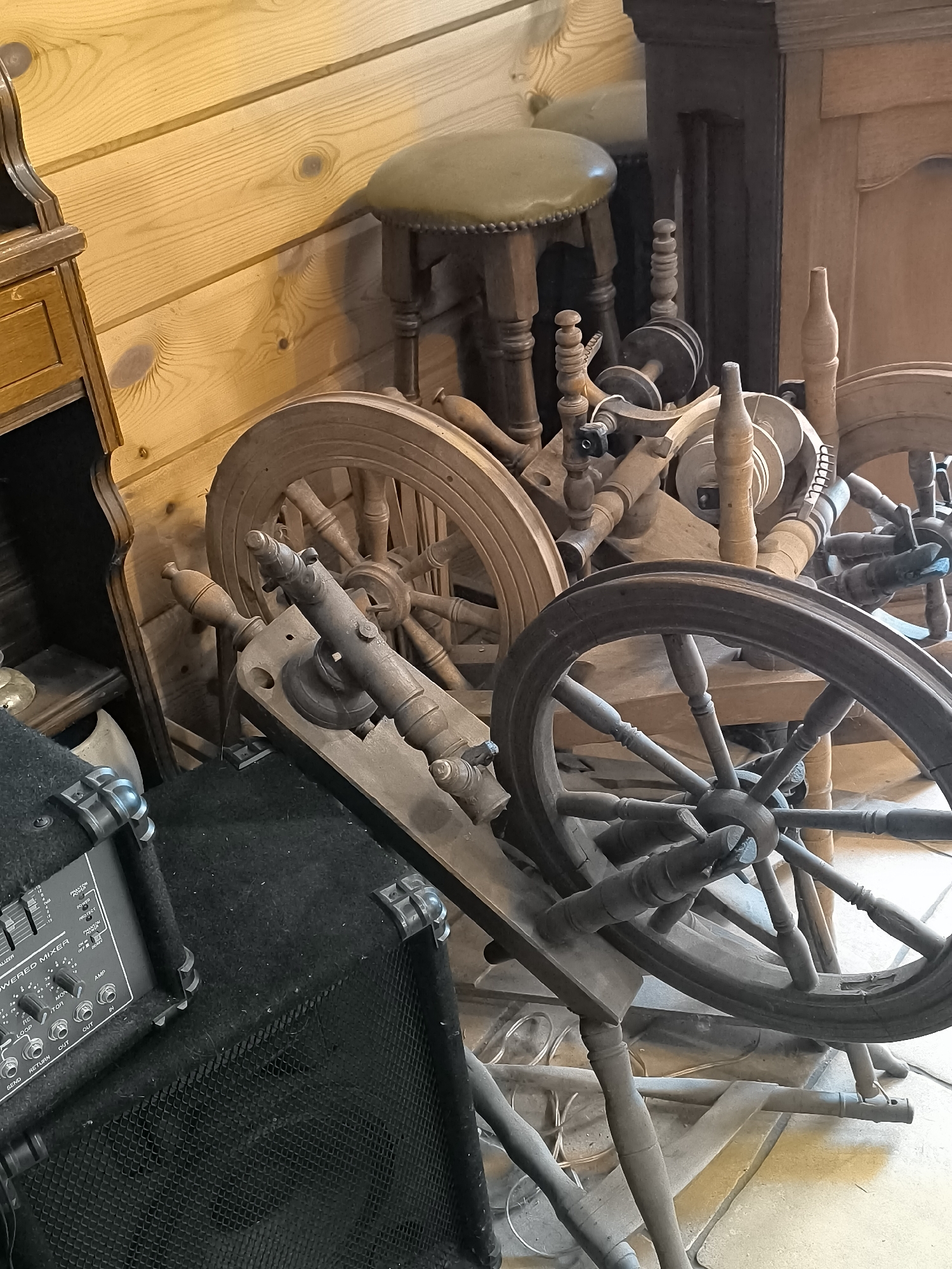 The image depicts an indoor scene with a collection of antique spinning wheels and other wooden furniture, including a stool. The walls are constructed of light-colored wooden planks. In the foreground, a black speaker or amplifier with control knobs and labels such as "POWERED MIXER," "MONITOR," "AMP," and "IN" is visible. The spinning wheels are made of dark wood and are intricately designed with large spoked wheels. Some of the wheels are stacked or positioned closely together, suggesting they are part of a collection or display. The overall impression is one of a storage area or a historical exhibit. The scene is brightly lit, possibly by natural light coming from a window or overhead lights. Based on the artistic style and subject matter, this image could potentially be located in Kuldīga, Latvia, a city known for its historical architecture and cultural heritage.