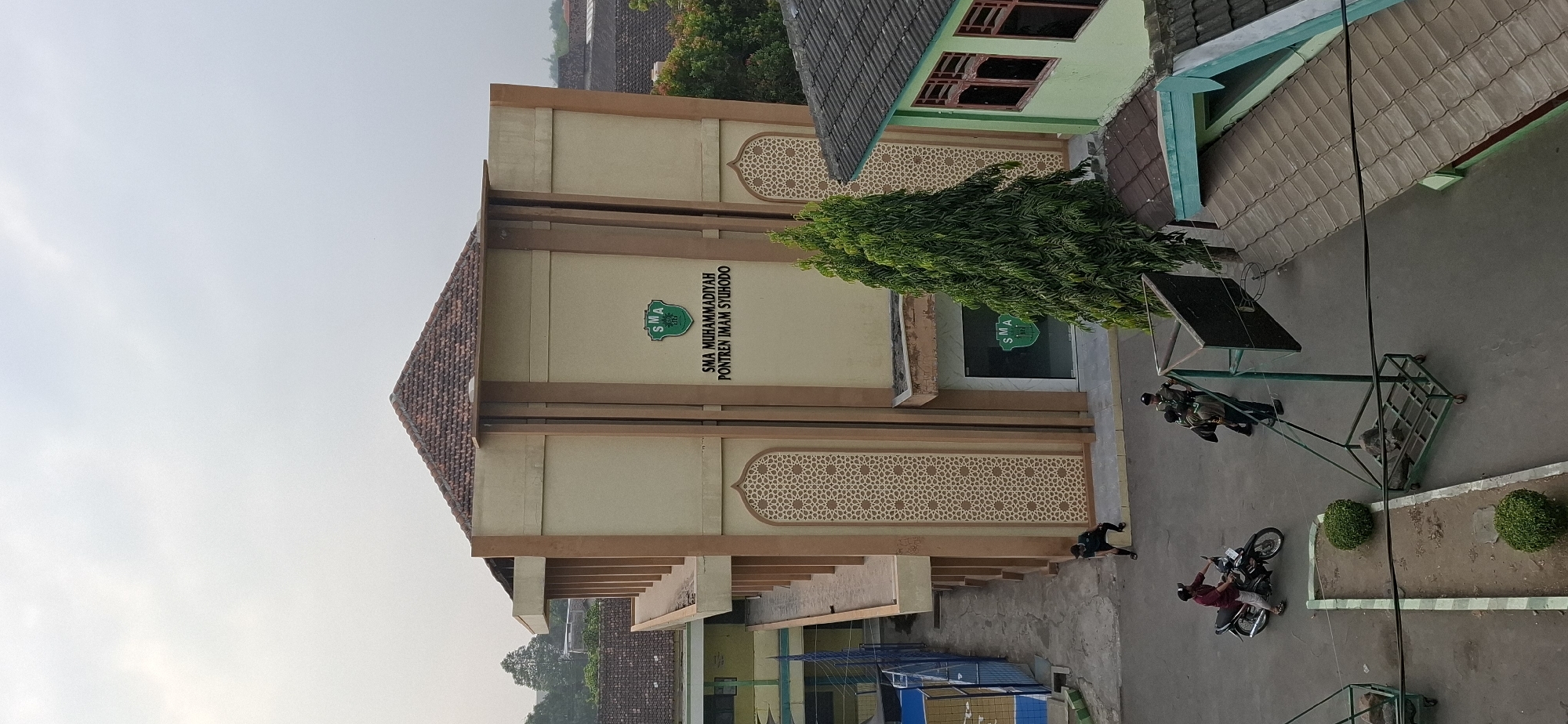 This is an outdoor, medium-angle shot of the SMA Muhammadiyah school building in Wonorejo, Indonesia. The building is a two-story structure with a beige facade, a brown tiled roof, and decorative Islamic-patterned panels. Above the entrance, the school's logo and name are displayed. In front of the building, a tall, slender green tree stands prominently.
On the paved ground in front of the building, there are several people and objects. A person in a maroon shirt and dark pants is riding a black motorcycle. To the right of the motorcycle, two individuals in military-style uniforms are standing next to a basketball hoop. Another person in dark clothing is sitting on a low wall near the side of the building.
The surrounding environment includes other buildings with tiled roofs, fences, and lush green trees, suggesting a suburban or semi-rural setting. The sky is overcast.