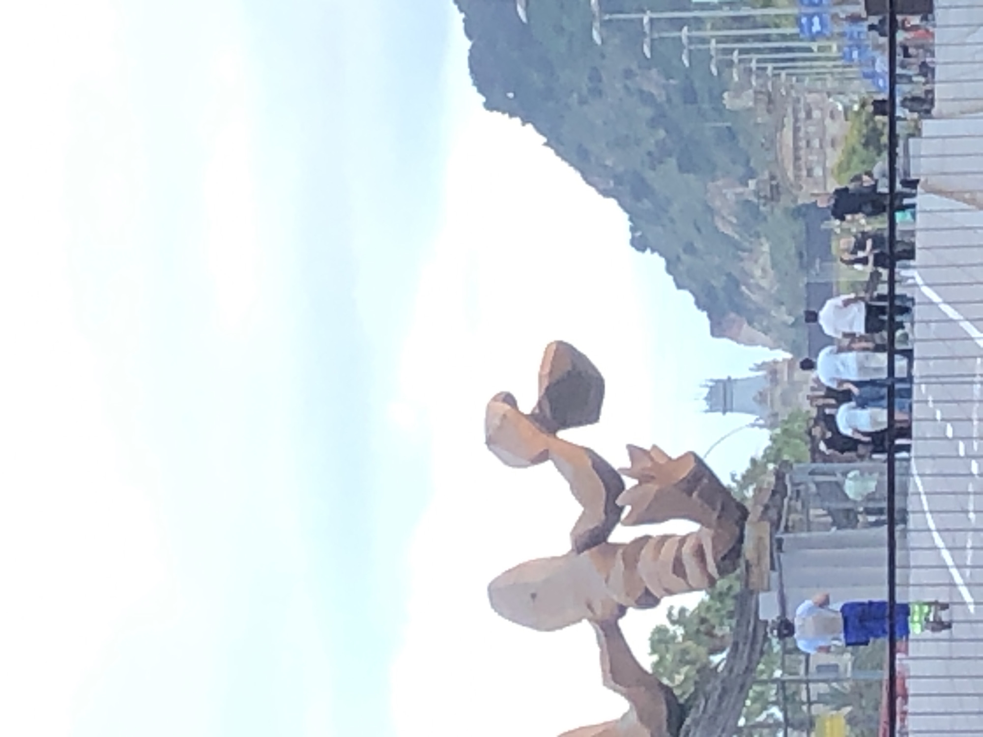 An outdoor, medium-shot image shows a sculpture of a large, abstract crab or lobster in the foreground. The sculpture is made of brown, segmented material and appears to be a landmark. In the background, there are several people walking and standing. To the right of the sculpture, a green, tree-covered mountain rises. Further in the background, there are buildings and streetlights. The sky is a pale blue with some clouds. One person on a scooter is visible in the lower left, wearing a blue shirt, a backpack, and brightly colored pants.