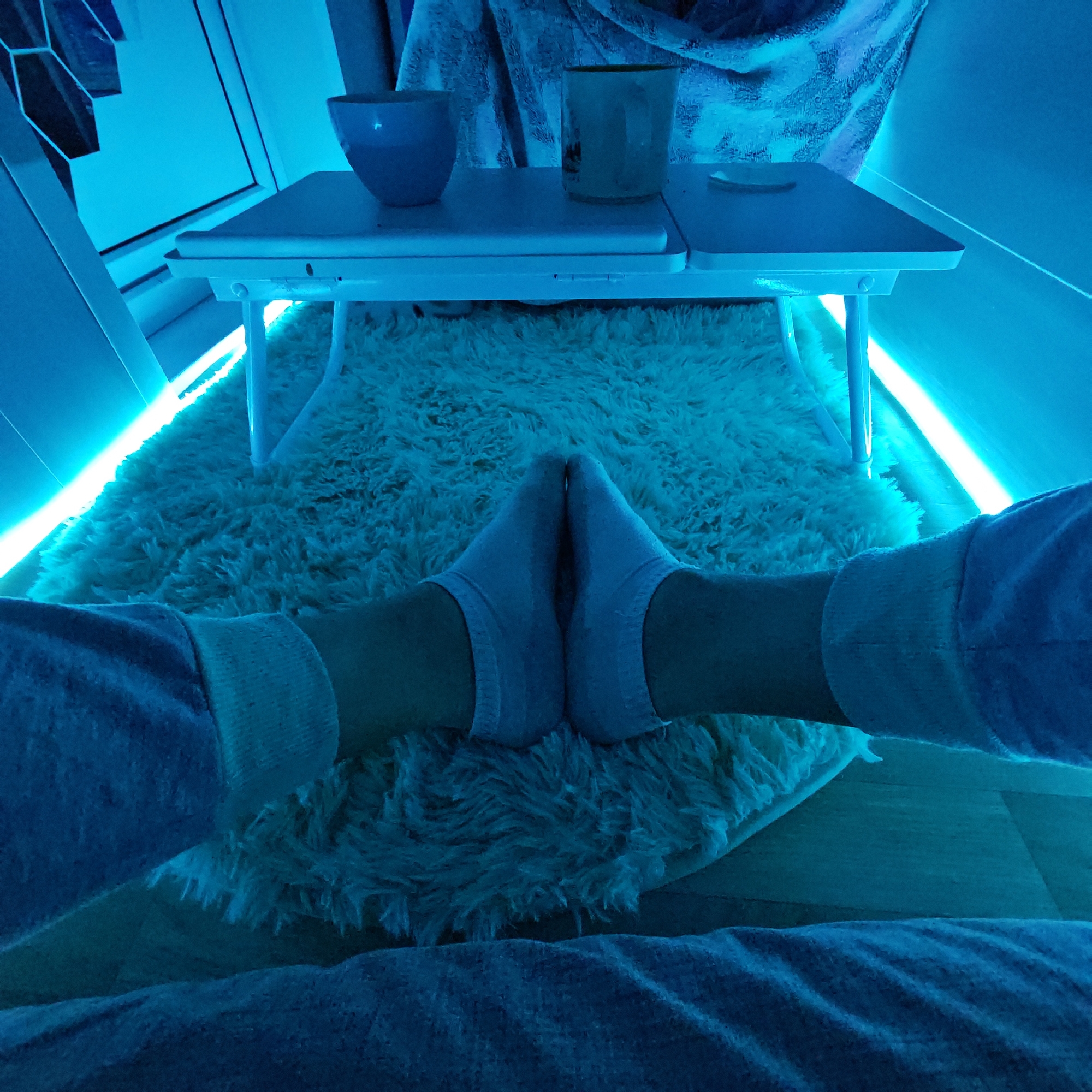 A first-person perspective photograph taken indoors. The viewer's legs, clad in light gray sweatpants and white socks, are visible in the foreground. The feet are pointed inward, nearly touching.
A small, white, foldable table rests on a white, shag rug. On the table are two cups, one blue and one clear.
LED strip lighting gives the scene a blue hue. Wood flooring is visible at the bottom of the frame. A window and wall are visible in the background, suggesting an indoor location.
There is no specific information provided about the location being Rostov, Russia.