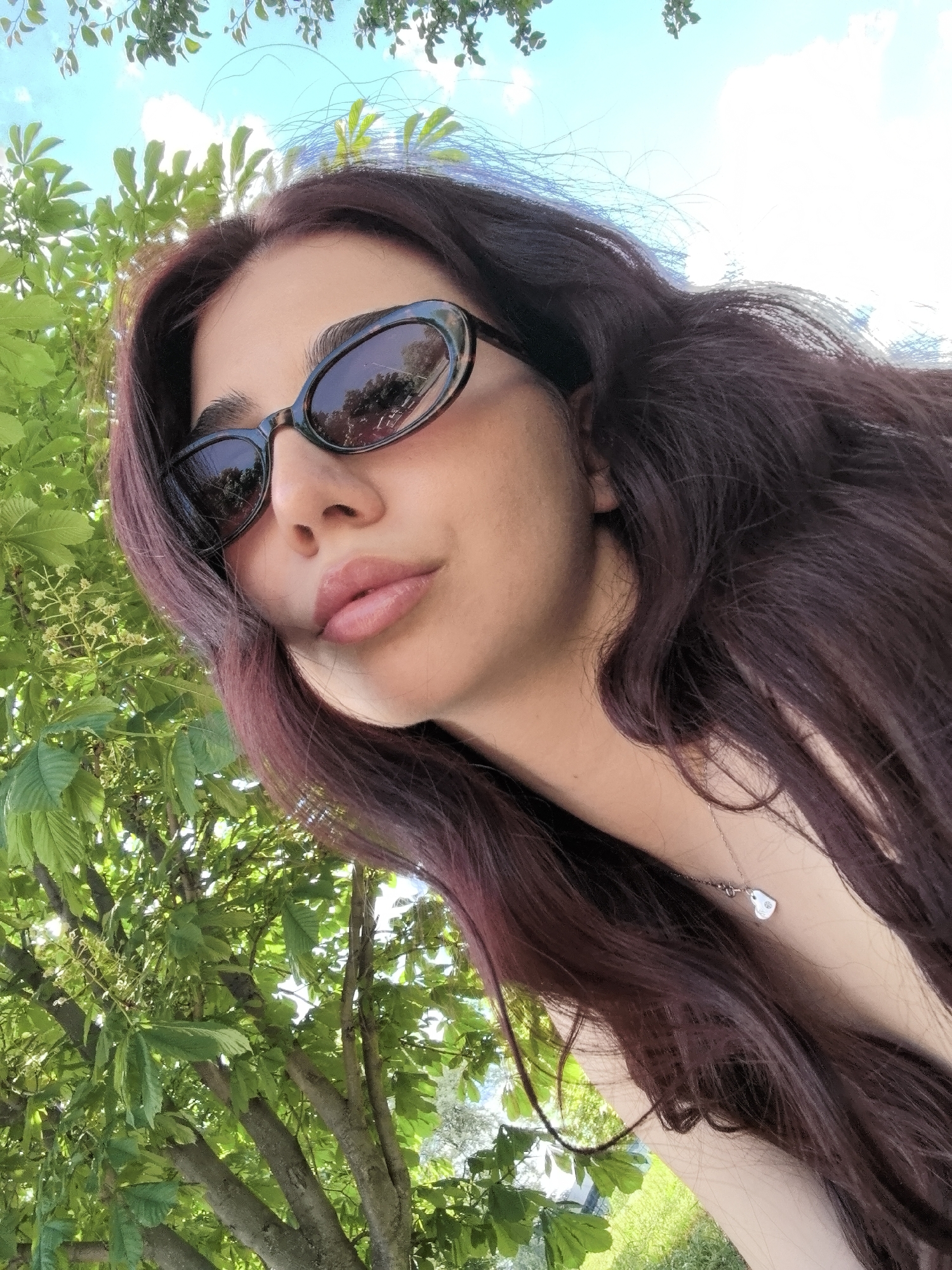 A woman with long, dark auburn hair is photographed from a low angle, positioned against a backdrop of trees and sky, likely in an outdoor setting. She is wearing dark sunglasses. A silver necklace with a small pendant is visible on her neck. The background reveals green foliage and branches of trees against a bright sky. The exact location within Bogomilovo, Bulgaria, cannot be determined with the given visual information.