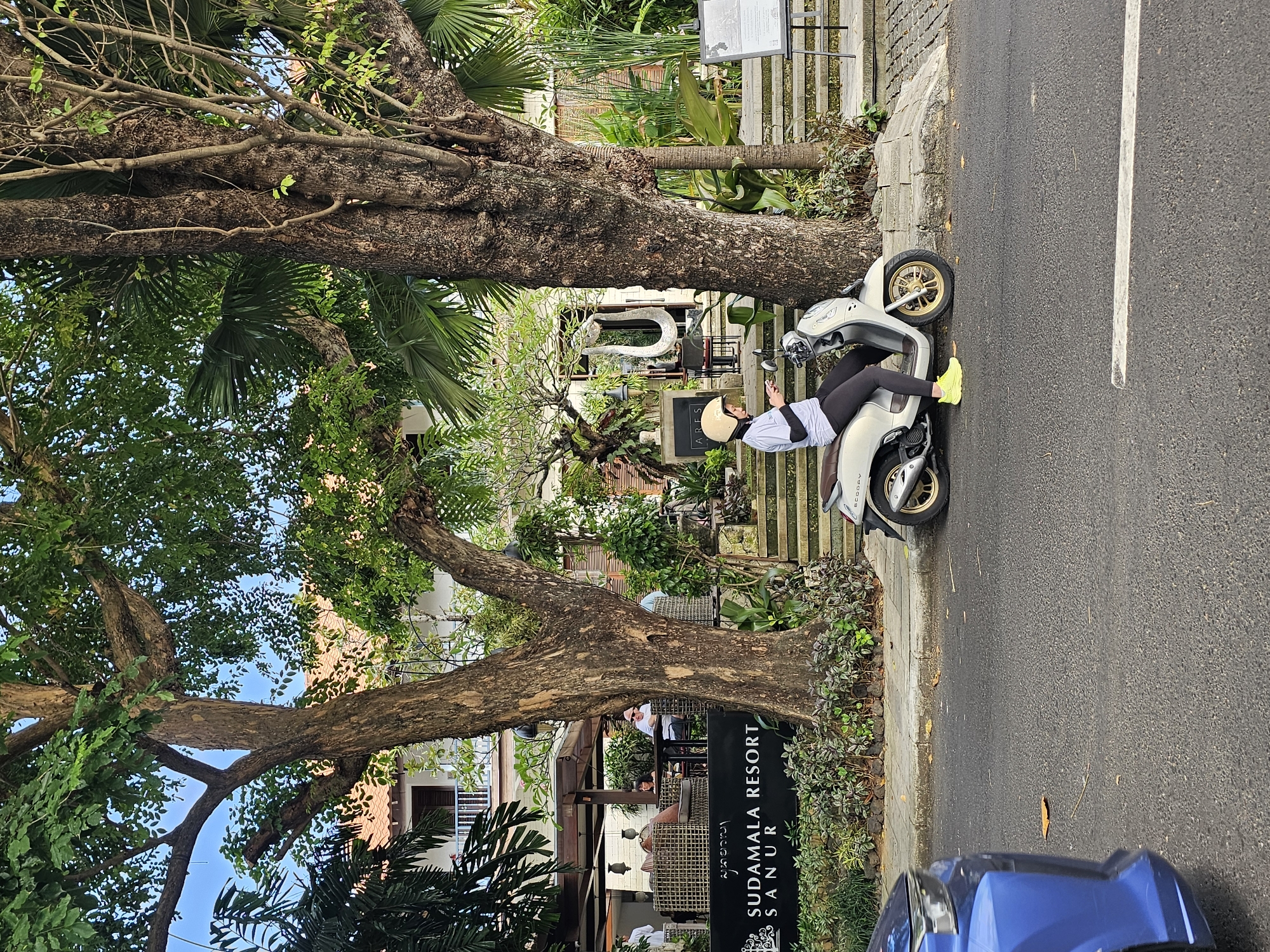 The image depicts a street scene in Denpasar, Indonesia. A woman, wearing a helmet and bright yellow sneakers, is seated on a silver scooter parked on the sidewalk. She appears to be using a phone. The scooter is parked next to a large tree and the entrance to the "Sudamala Resort Sanur". A blue car is partially visible at the bottom of the frame. The scene includes trees, foliage, and a paved road, suggesting an urban setting.