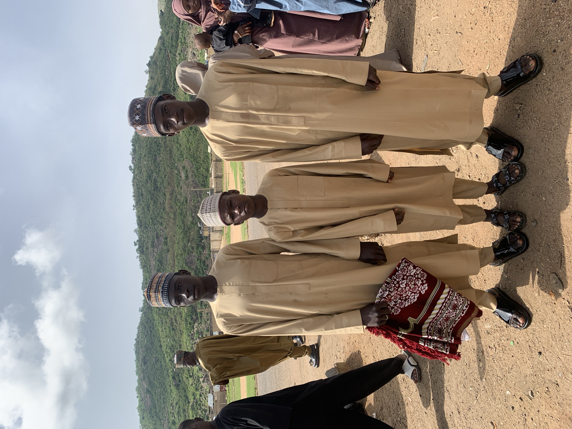 The photo depicts three young men standing on a paved road in Naraguta Hausawa, Nigeria. They are dressed in tan-colored, traditional garments and hats. The man in the bottom of the photo holds a folded, red and white prayer rug. They are all wearing sandals. Behind them is a hillside covered in green vegetation under a cloudy sky. Additional people are visible in the background wearing various colors of dresses and hijabs. Another individual is visible in the bottom of the photo wearing a black robe.