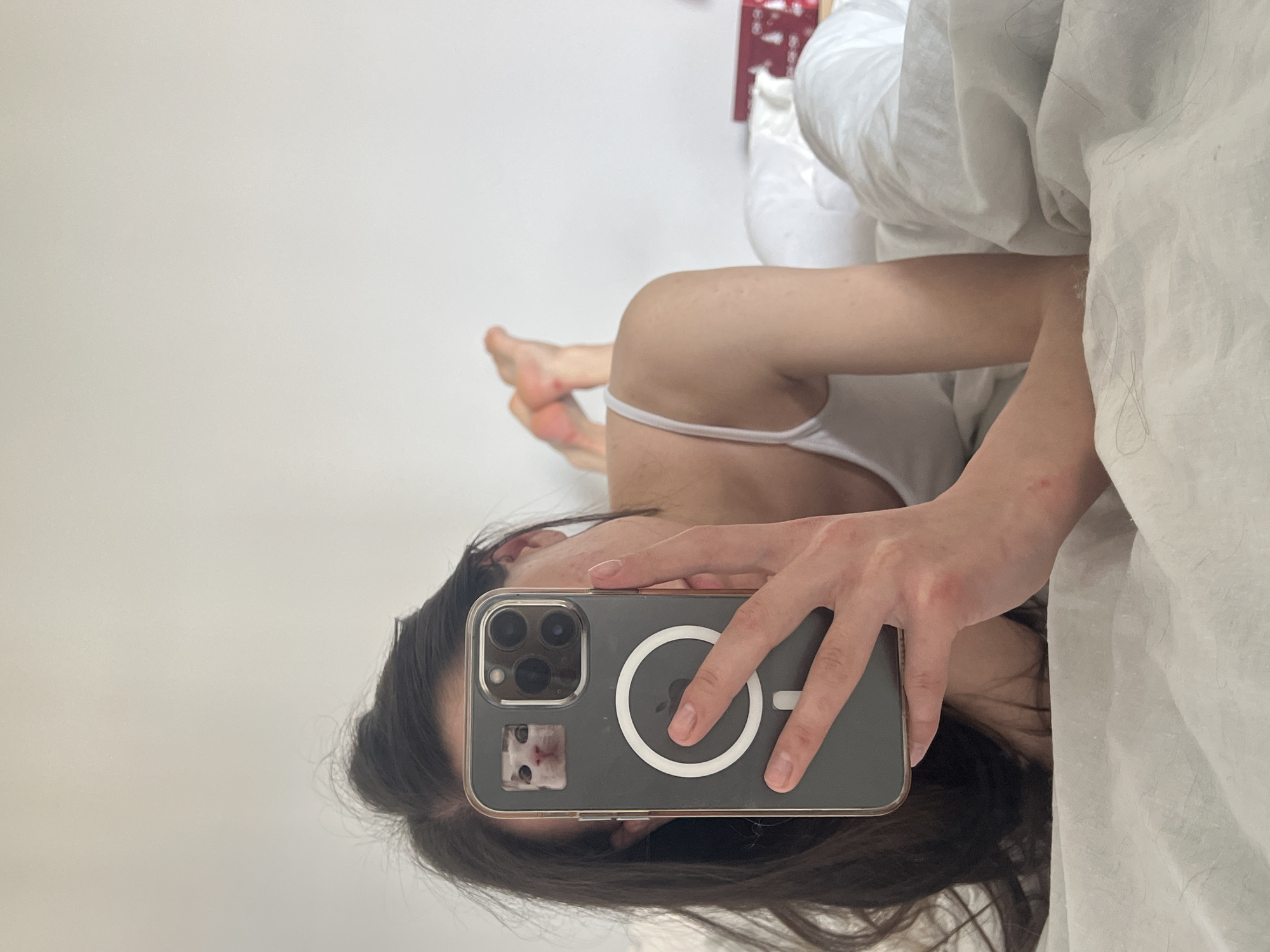 A woman is lying in bed taking a selfie with a smartphone in a clear case. The phone case has a white circle attachment and a photo of a white cat. The woman is wearing a white tank top and is covered by white bedding. Her feet are visible in the background. The location is not specified and cannot be determined from the image.