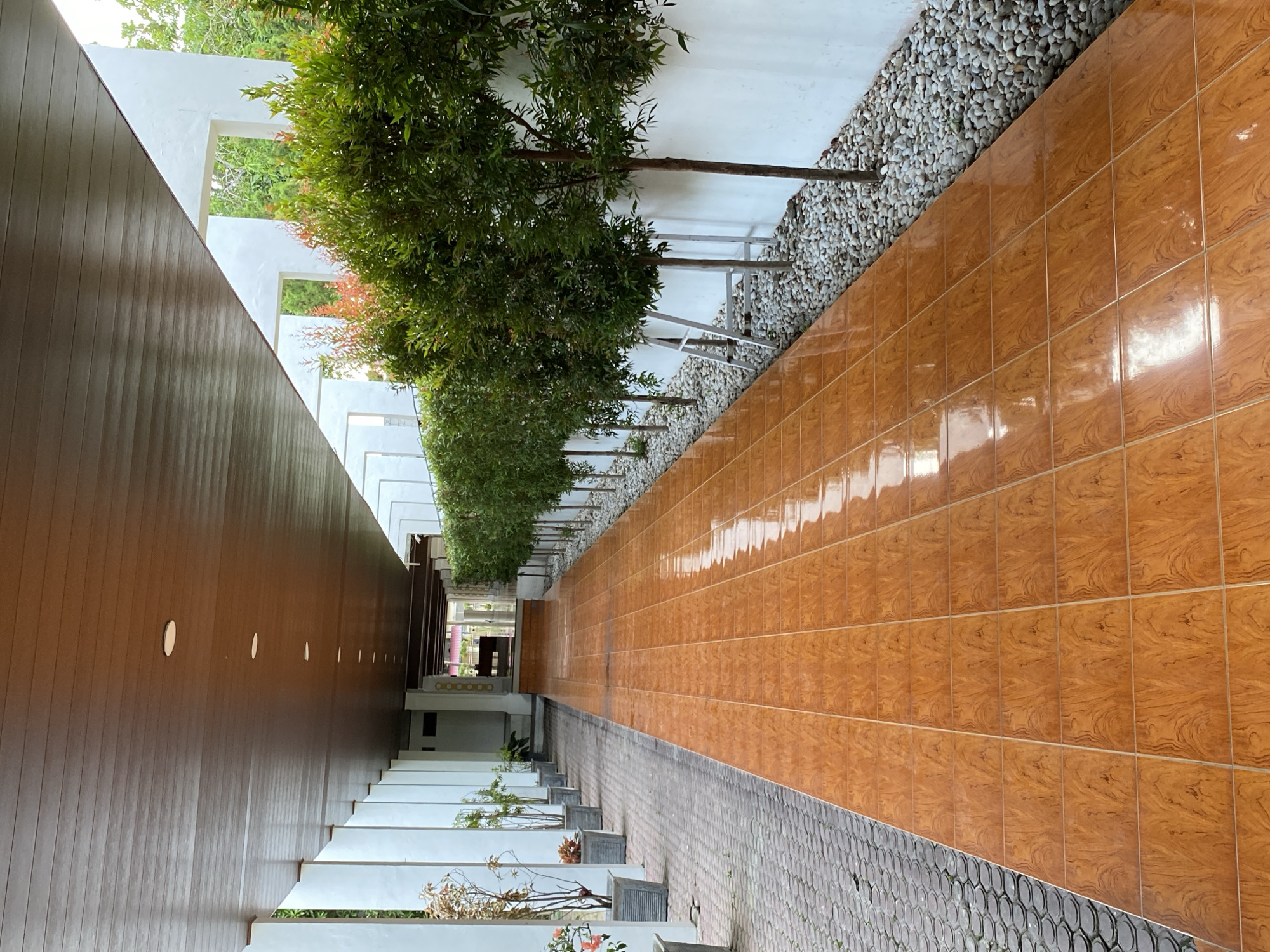 This is an outdoor image of a covered walkway in Banda Aceh, Indonesia. The walkway is lined with white pillars on the left and small trees on the right, separated by a wall. The ground is a combination of patterned grey pavers on the left and glossy orange tiles on the right. The orange tiles reflect the overhead lights. The walkway leads towards an open doorway in the distance, suggesting an entrance to a building or complex. The overall scene depicts a clean and well-maintained outdoor passage.