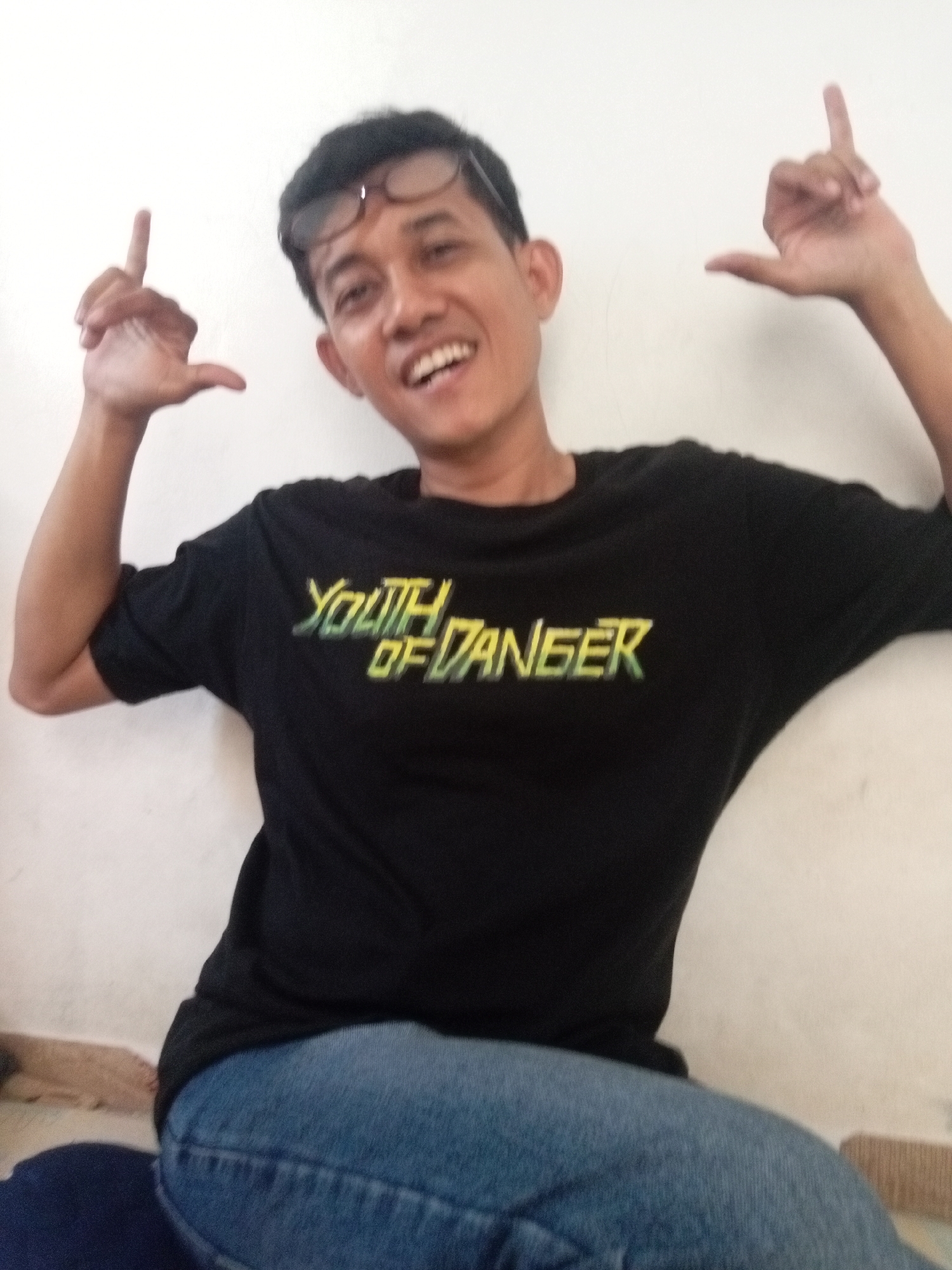 The image shows a medium shot of a young adult male with black hair, wearing glasses, a black t-shirt with yellow text that reads "YOUTH OF DANGER", and blue jeans. He is posing with his hands up, fingers pointing towards the camera, and a wide smile. The background appears to be a plain, light-colored wall. The overall scene suggests an informal indoor setting. The image quality is somewhat blurred, particularly around the edges and the subject's facial features. There is no visual information within the image that indicates the location as being in the city of Batam, Indonesia.