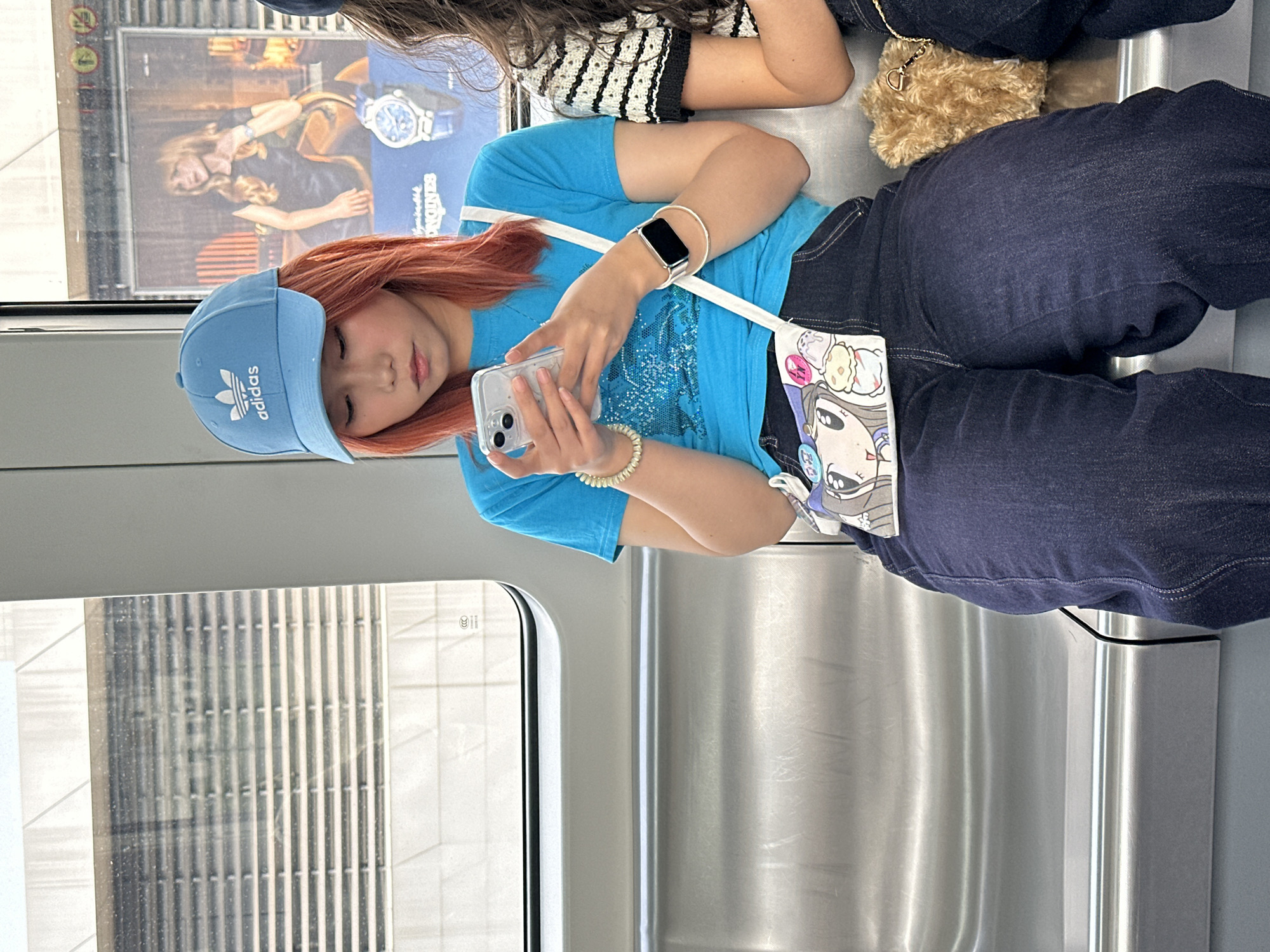 A young person wearing an Adidas baseball cap, a blue t-shirt, and jeans is sitting on a train or bus. They are looking at and using a phone. A building is visible through the window. A Longines ad is on the wall. This appears to be in the Jiulongpo District, China, based on the ad.