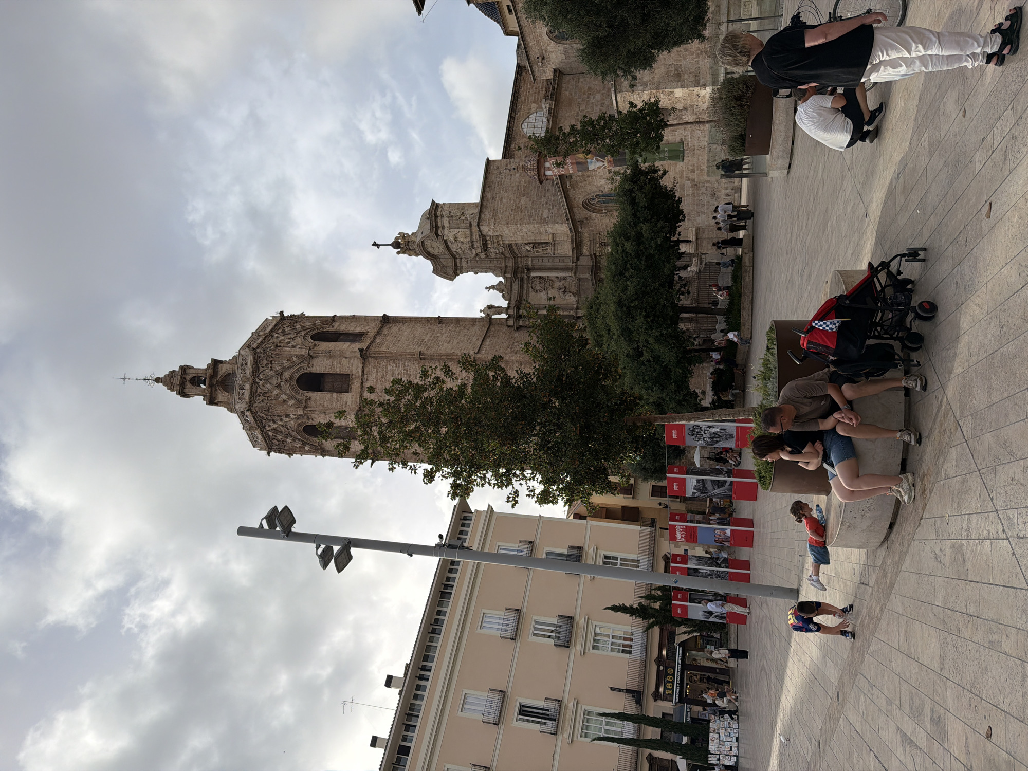 The image depicts a street scene in front of a large, historic stone church with a prominent bell tower. The church has intricate architectural details and is surrounded by trees. To the left of the church is a light-colored, multi-story building with balconies and windows. The ground is paved with light-colored square tiles, and a circular area with a bench is visible in the foreground.
Several people are present in the scene. A family of three, two adults and a child, are sitting on the circular bench. The adults are facing the church, and the child is leaning on the bench. Another child in a blue and yellow shirt is on the ground in front of the bench, appearing to be picking something up. Further to the right, a person in a black shirt and white pants is standing near a bicycle. Another person in a white shirt is crouching down near the bicycle. In the background, more people are visible walking and sitting in front of the buildings and church. Several red exhibition stands are set up in the plaza, displaying photographs. The sky is overcast with clouds. The overall impression is of a lively public square in a European city.