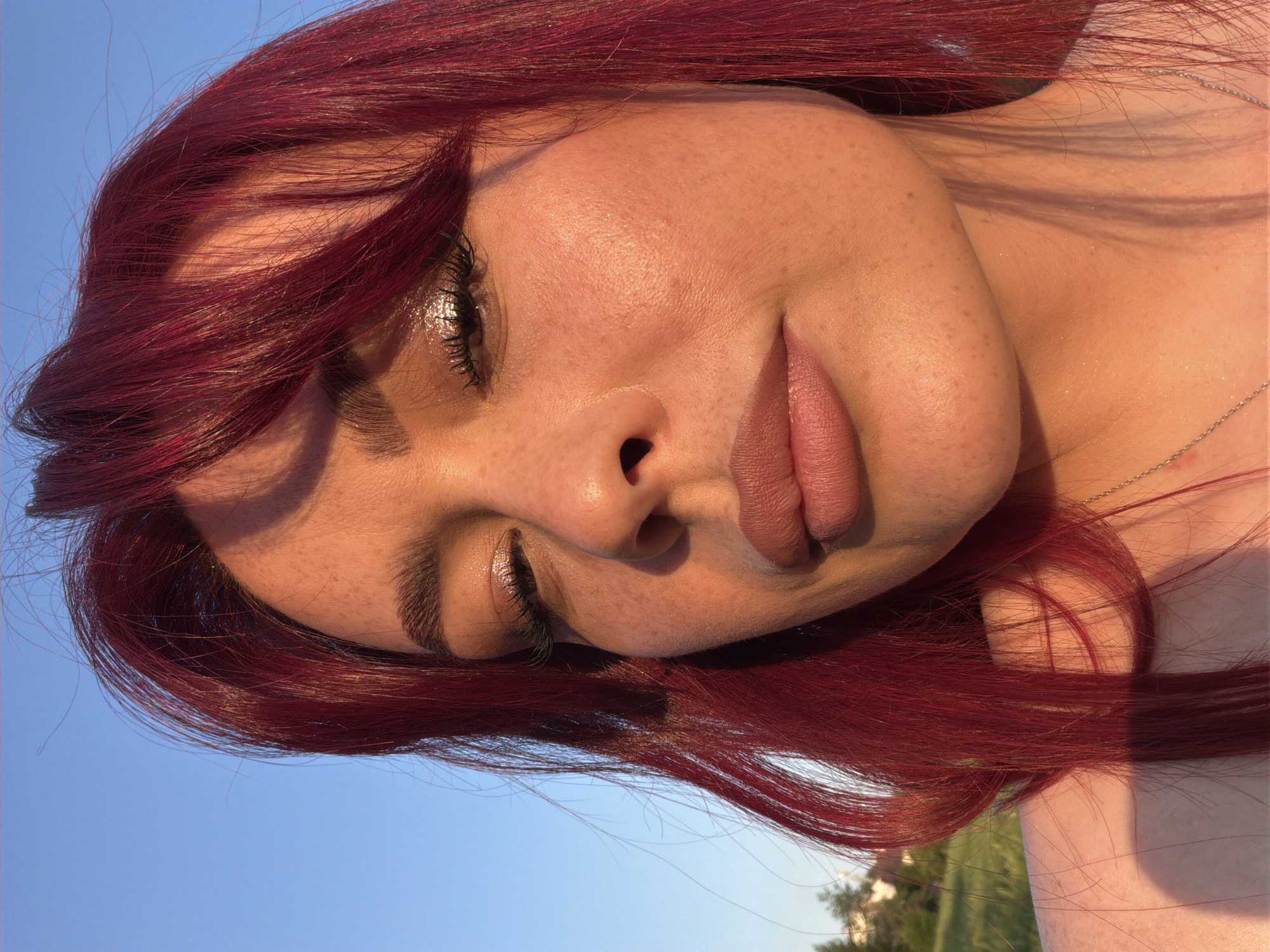 A close-up, eye-level, outdoor portrait captures a young woman with bright red hair against a clear blue sky during golden hour. The sunlight casts a warm glow on her face, highlighting her freckles, makeup, and the shimmer on her eyelids. Her eyes are partially closed, and she looks toward the camera. The background shows a blurred green field and trees, suggesting a park or natural area within Tolyatti, Russia.