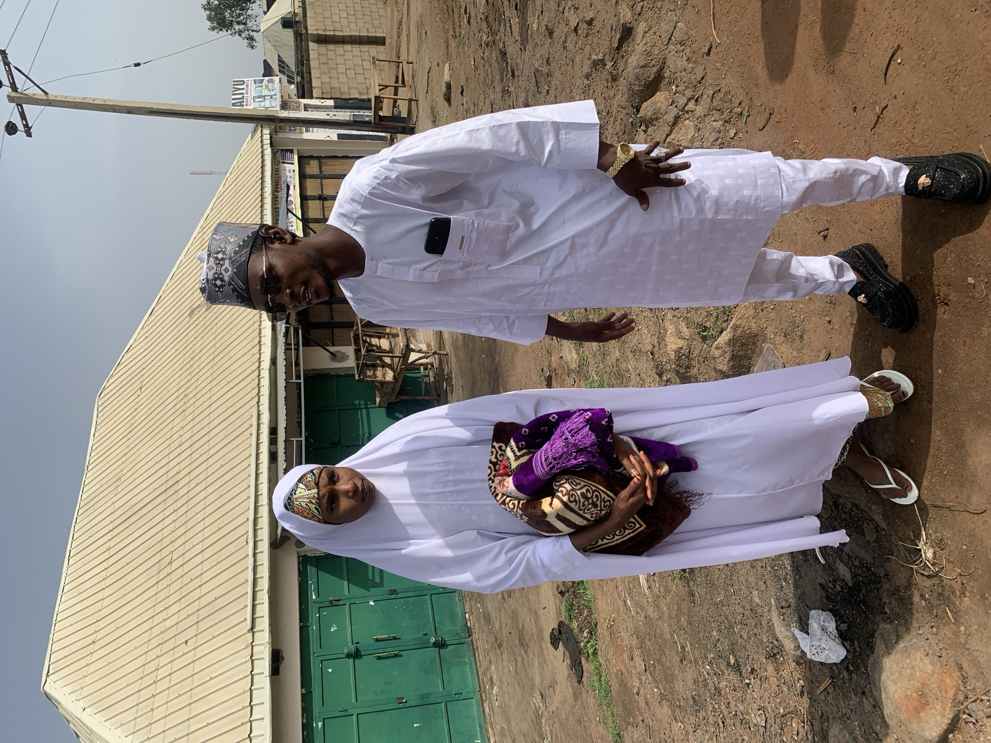 Two individuals are centrally positioned in an outdoor setting, likely Naraguta Hausawa, Nigeria. A man, wearing a white tunic and trousers, a decorative hat, and sunglasses, stands with his right hand raised. A dark-colored smartphone is visible in his shirt pocket. He is wearing black shoes with gold embellishments. Next to him, a woman is dressed in white clothing, holding a prayer mat with purple and brown patterns. She also wears decorative headwear. They are standing in front of a building with green doors. The ground appears to be dirt or gravel. A power pole with attached utility lines is visible. Another building with a sign that reads "ALIYU...ABILITY BUSINESS" is in the background.