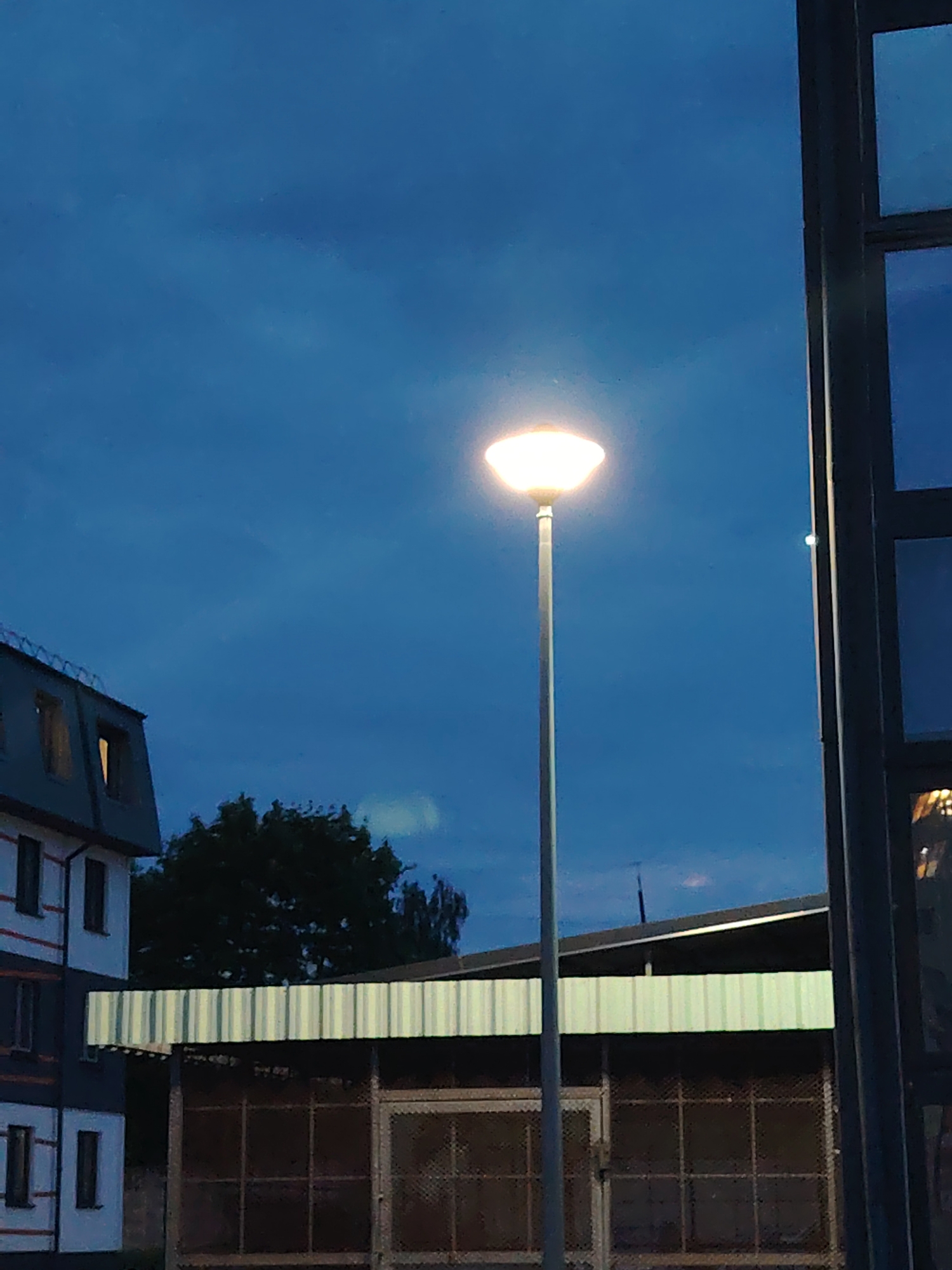 The image captures an outdoor scene at dusk, likely in the Бараўлянскі сельскі Савет, Belarus, featuring a lit streetlamp in the foreground. The sky is a deep blue, indicative of twilight, with some faint cloud formations. To the left of the streetlamp, a multi-story building with a dark, angled roof and white and dark gray siding is visible. Several windows with yellowish light emanating from within are present on this building. Behind the building and to the right of the streetlamp, a dense cluster of dark green trees stands out. In the middle ground, a building with a corrugated metal roof, striped in alternating white and light gray, spans horizontally across the frame. Beneath this roof, a structure with a dark, grid-like facade, possibly a fence or enclosure, is visible. To the far right of the image, a dark vertical element, appearing to be part of a window frame or building structure, partially obscures the view. The overall atmosphere suggests a quiet, residential or semi-industrial area at the transition from day to night.