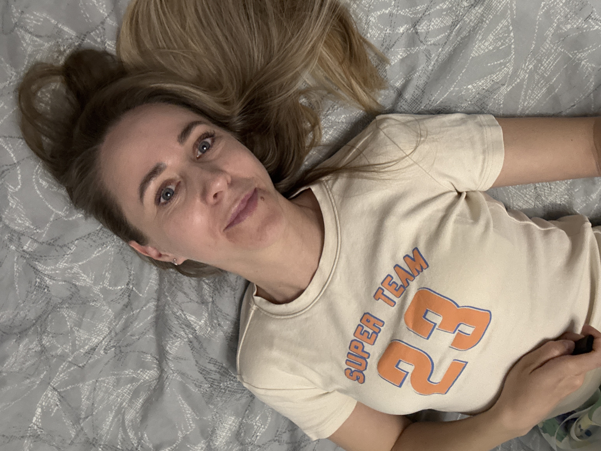 A blonde woman lies on her back on a grey patterned bedspread. She wears a beige T-shirt with "SUPER TEAM 23" printed in orange and blue. Her hair is spread around her head on the bedspread. The location cannot be confirmed as Chaikovsky, Russia, based on the provided image.