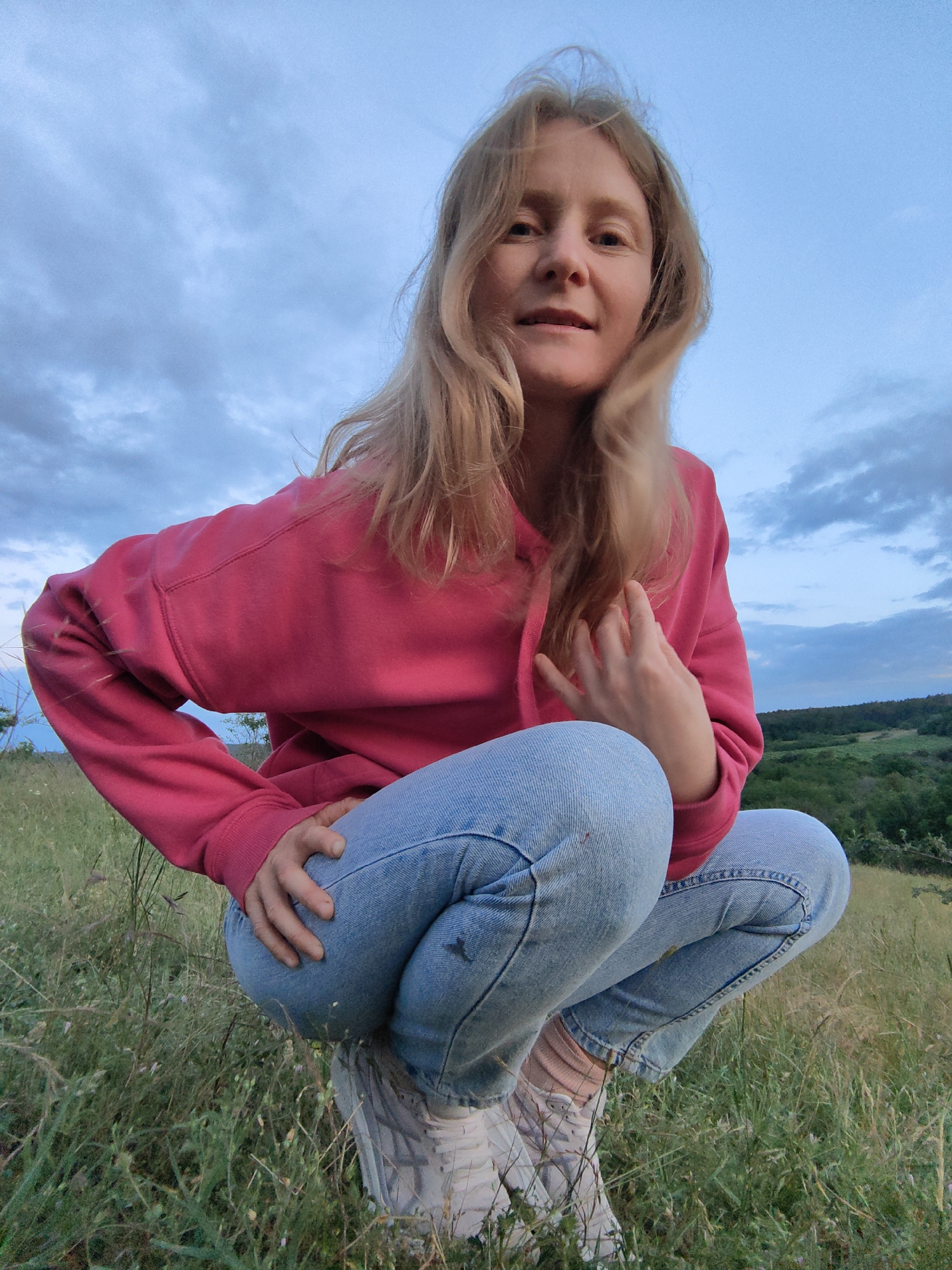 A woman with blonde hair is crouching in a grassy field. She is wearing a pink hooded sweatshirt and light-wash jeans. Her hair is flowing around her face, and she is looking at the camera. The background shows a slightly cloudy sky and a green, rolling landscape. The image appears to have been taken outdoors during either dusk or dawn, with soft lighting. The setting is natural and somewhat wild, with tall grass and vegetation visible in the foreground. The woman's posture and gaze suggest a relaxed and candid moment. There is no visible text in the image.