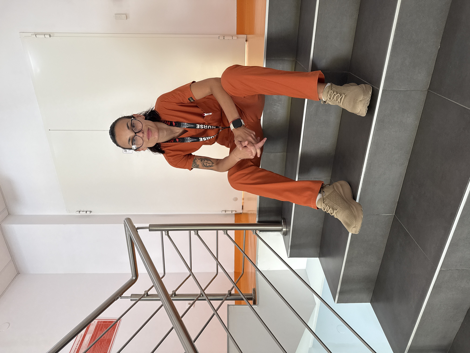 The image depicts a female individual seated on a dark grey tiled staircase within a modern building in Pleven, Bulgaria. She has dark hair pulled back, wears dark-framed eyeglasses, and looks directly at the camera with a neutral expression and slight smile.
The individual is dressed in an orange V-neck scrub top and matching pants, indicative of medical professional attire. A black lanyard with white "NURSE" text and a medical cross emblem hangs around her neck, attached to a clear ID holder. A white smartwatch with a red band is worn on her left wrist, and a prominent tattoo depicting faces is visible on her left upper arm/bicep. Her hands are clasped in her lap, and she wears light brown athletic shoes with thick soles. She is positioned on the second step from the bottom of the staircase, with her legs extended forward.
The staircase features dark grey treads and risers, each tread edged with a thin silver metallic strip. A stainless steel railing with multiple horizontal bars runs alongside the stairs. The surrounding walls are predominantly white. In the background, an orange-colored floor is visible on the upper level. To the left, a red rectangular sign with white Cyrillic text, partially legible as "СЪРЦЕ И МОЗЪК СЕВЕР" (HEART AND BRAIN NORTH), is mounted on the white wall next to the railing. To the right, a large white door with visible hinges is present, and a white electrical outlet or switch is affixed to the wall further right. The ceiling appears to be a white suspended panel system.