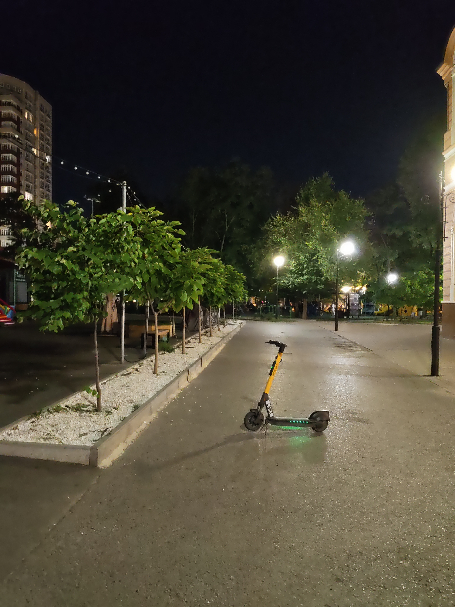 This image depicts an evening scene in Rostov-on-Don, Russia, likely in a park or public square. The foreground is dominated by a wet asphalt path where a yellow and black electric scooter is parked. The scooter has green lights illuminated on its base. To the left of the path, a row of young, leafy trees are planted in a raised bed filled with white gravel. Beyond the trees, a large, multi-story residential building with illuminated windows is visible against a dark night sky. Several streetlights are present throughout the scene, casting a warm glow on the surroundings. In the background, more trees and what appear to be playground structures are visible, suggesting a recreational area. The overall impression is one of a quiet urban park at night.