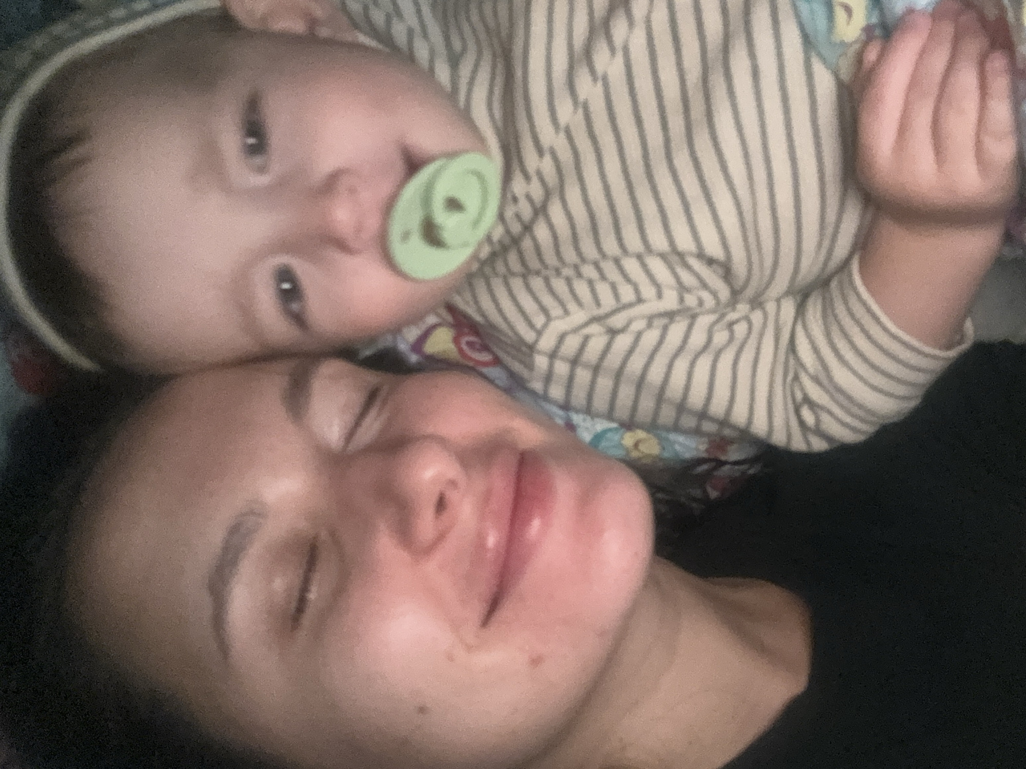 A photograph shows a woman and a baby laying on a bed or surface. The woman is on the bottom, and the baby is resting on top. The woman is lying on her back with her eyes closed and a relaxed expression. The baby is wearing a striped outfit and has a green pacifier in its mouth. The baby is facing the camera with eyes open. The location of the image is unknown.