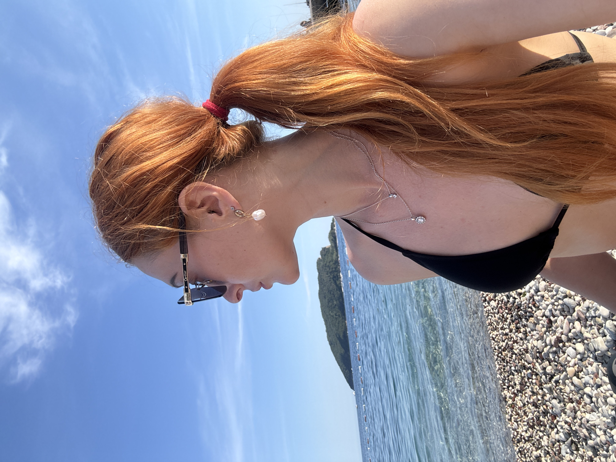 A red-haired woman with sunglasses is seen from the side, standing on a pebble beach next to the Adriatic Sea in Budva, Montenegro. She is wearing a black bikini and a necklace. Her hair is pulled back in a ponytail. The sky is blue with some clouds, and there's a tree-covered island in the background.