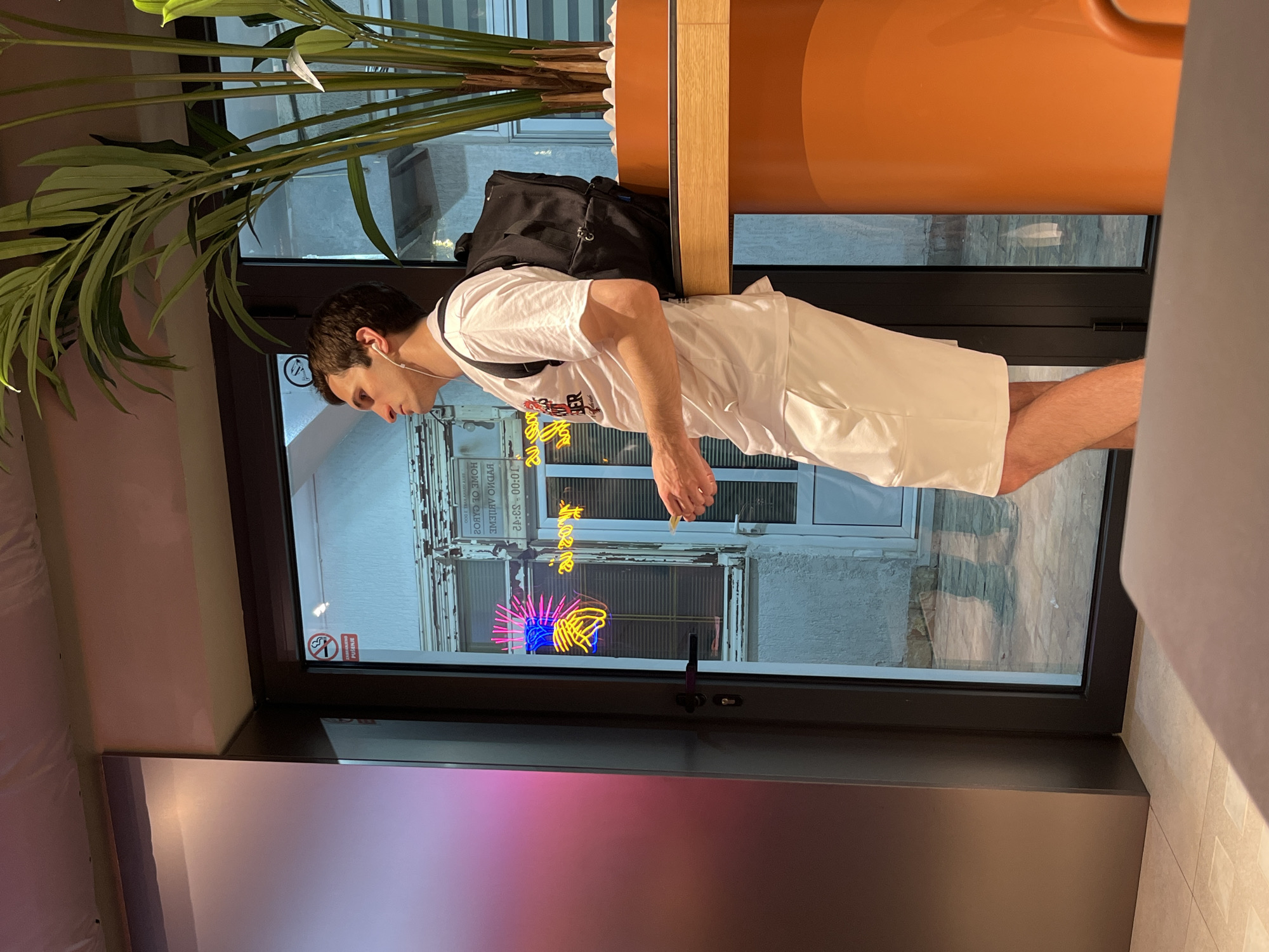A young man wearing a white t-shirt, white shorts, and a black backpack stands near a glass door in what appears to be an establishment in Budva, Montenegro. He is looking to his left, with white earbuds in his ears. Through the glass door, neon lights depicting hands are visible, as is a "No Smoking" sign. A large potted plant with green fronds stands to the right of the man, next to an orange cylindrical object. The floor appears to be tiled.