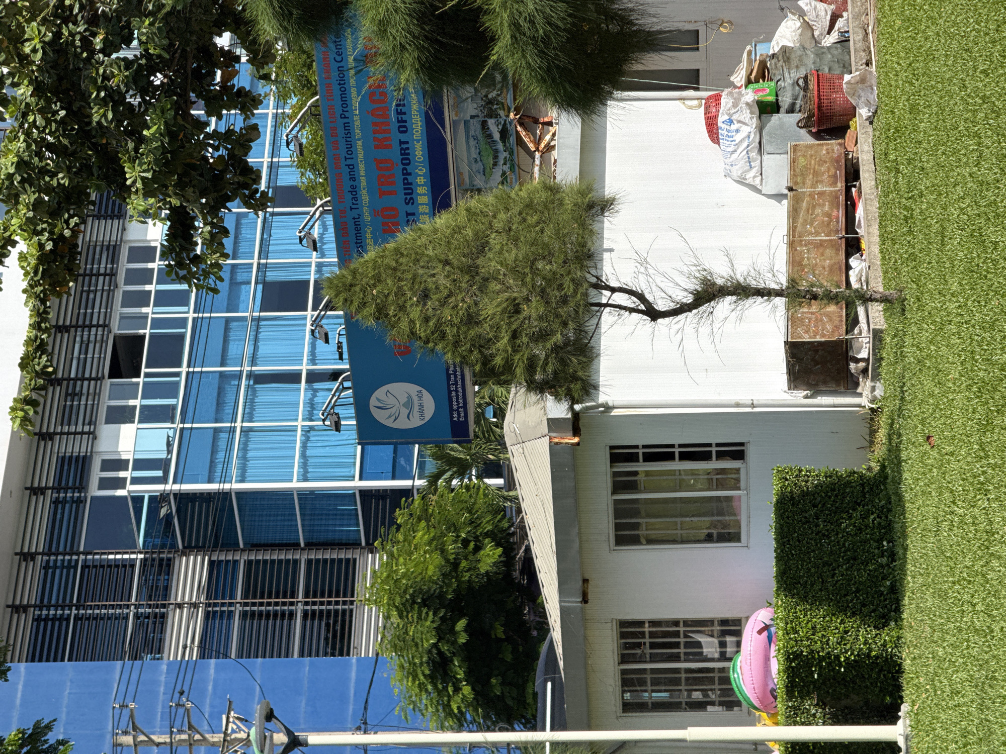 The image depicts a street scene in Nha Trang, Vietnam, with a modern building in the background and a smaller, temporary structure in the foreground. The building, likely an office or commercial establishment, features a facade dominated by large blue glass windows and horizontal metal louvers. In front of this building is a small, white, utilitarian structure with windows. To the left of this structure, a bright pink inflatable ring rests on a manicured green hedge.
The area in front of the smaller structure is cluttered with various items, including large sacks of material, red plastic baskets, and what appears to be a metal cage or cart. A thin, sparse tree stands in front of this clutter. A neatly mowed green lawn occupies the lower portion of the image. The overall impression is one of contrast between the polished modern architecture and the more ad-hoc arrangement of objects in the foreground, typical of many urban settings in Vietnam. The specific location within Nha Trang is not discernible from the image alone.