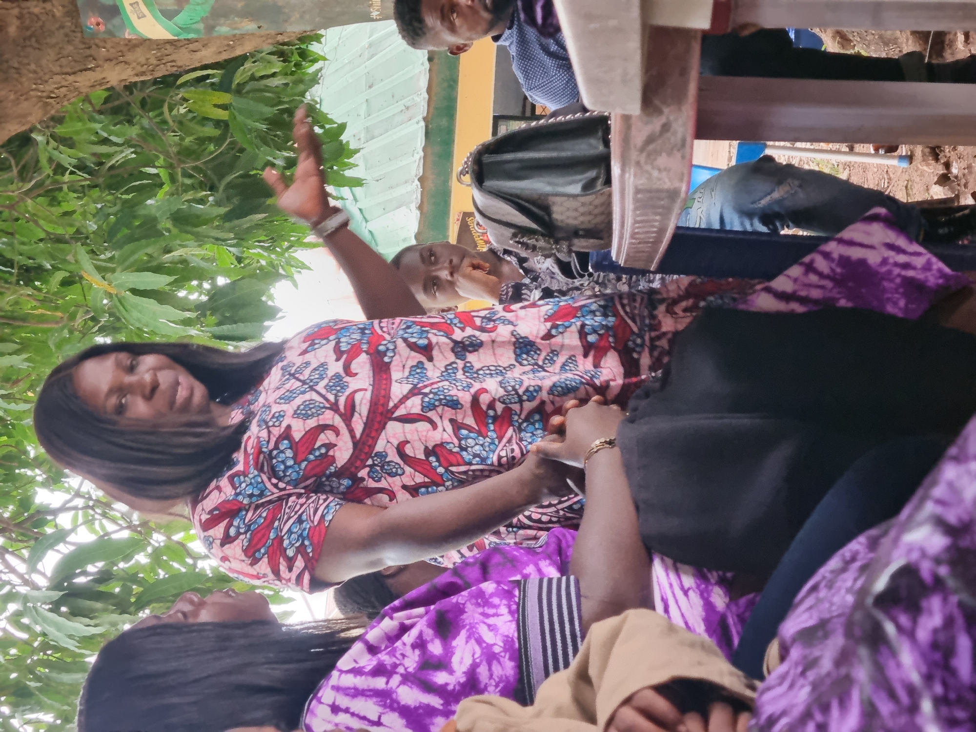 A group of people are gathered outdoors in what appears to be a covered or shaded area. The location is indicated as Jos, Nigeria.
A woman wearing a patterned pink, red, and blue top is the focal point. She is gesturing with her right hand raised. Other people are seated around her. One person wears a purple tie-dye patterned garment. A man is visible in the background wearing a blue shirt. A purse or bag is resting on a chair behind the woman. Vegetation, possibly trees, is visible in the upper left corner. A roof and wall are also visible in the background.