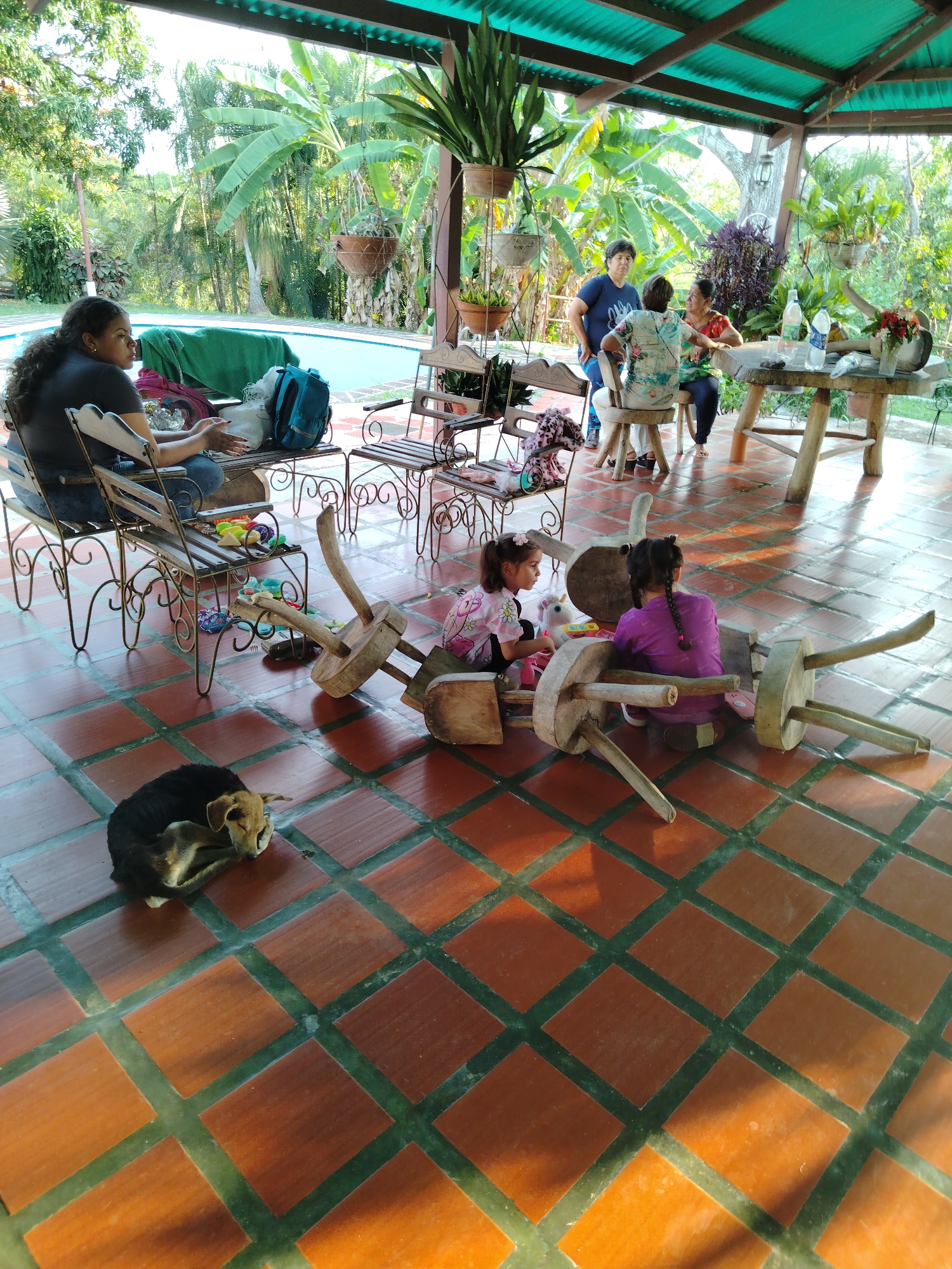 The image depicts an outdoor scene, possibly a patio or covered porch, in Los Leoncitos, Venezuela. The flooring consists of square terracotta tiles separated by green grout lines. The area is sheltered by a green metal roof.
Several individuals are present: a woman with dark curly hair is seated on a wrought iron bench to the left, looking towards the background. A group of three women are conversing at a rustic wooden table in the background. Two young girls are seated on the floor, playing with overturned wooden stools. A small dog lies curled up on the floor near the woman on the bench.
Other objects include potted plants, various chairs and benches of wrought iron and wood, a blue backpack, and a swimming pool visible in the background. The surrounding environment includes lush tropical vegetation.