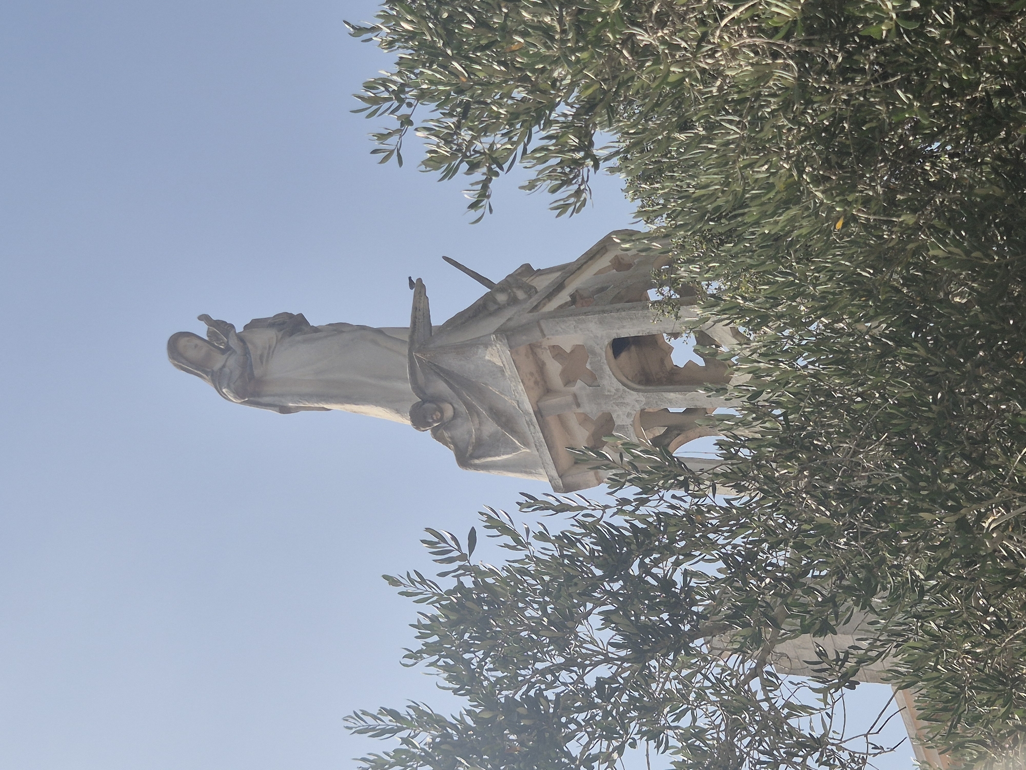 The image shows a statue of Mary and the infant Jesus atop a stone structure, partially obscured by olive tree branches. The statue is made of a light-colored material, possibly stone or concrete, and depicts Mary with a veiled head, holding a baby. The structure below the statue appears to be a bell tower or a similar architectural element, with arched openings and decorative stonework. A small bird is perched on the edge of the structure near the statue. The background is a clear blue sky, suggesting an outdoor setting. The presence of olive trees indicates a Mediterranean climate. This statue is located in the city of Abu Gosh, Israel, a town known for its Arab Christian community and religious sites.