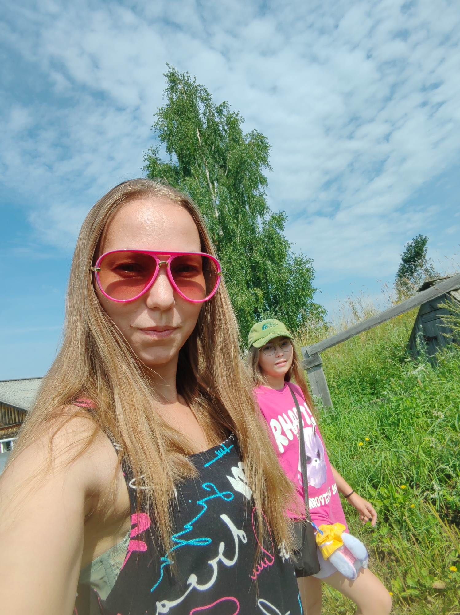 This is a selfie taken outdoors in a rural setting, likely in the city of Chukhlem, Russia, given the context. A woman with long blonde hair and large pink sunglasses is in the foreground, looking at the camera. She is wearing a black top with a colorful design. To her right and slightly behind her is a younger girl wearing a green cap and a bright pink t-shirt with a graphic print. She is also looking towards the camera. They are standing in a grassy area with tall trees and a rustic wooden structure in the background. The sky is blue with scattered white clouds, indicating a sunny day. The overall impression is one of a casual outdoor outing in a natural environment.