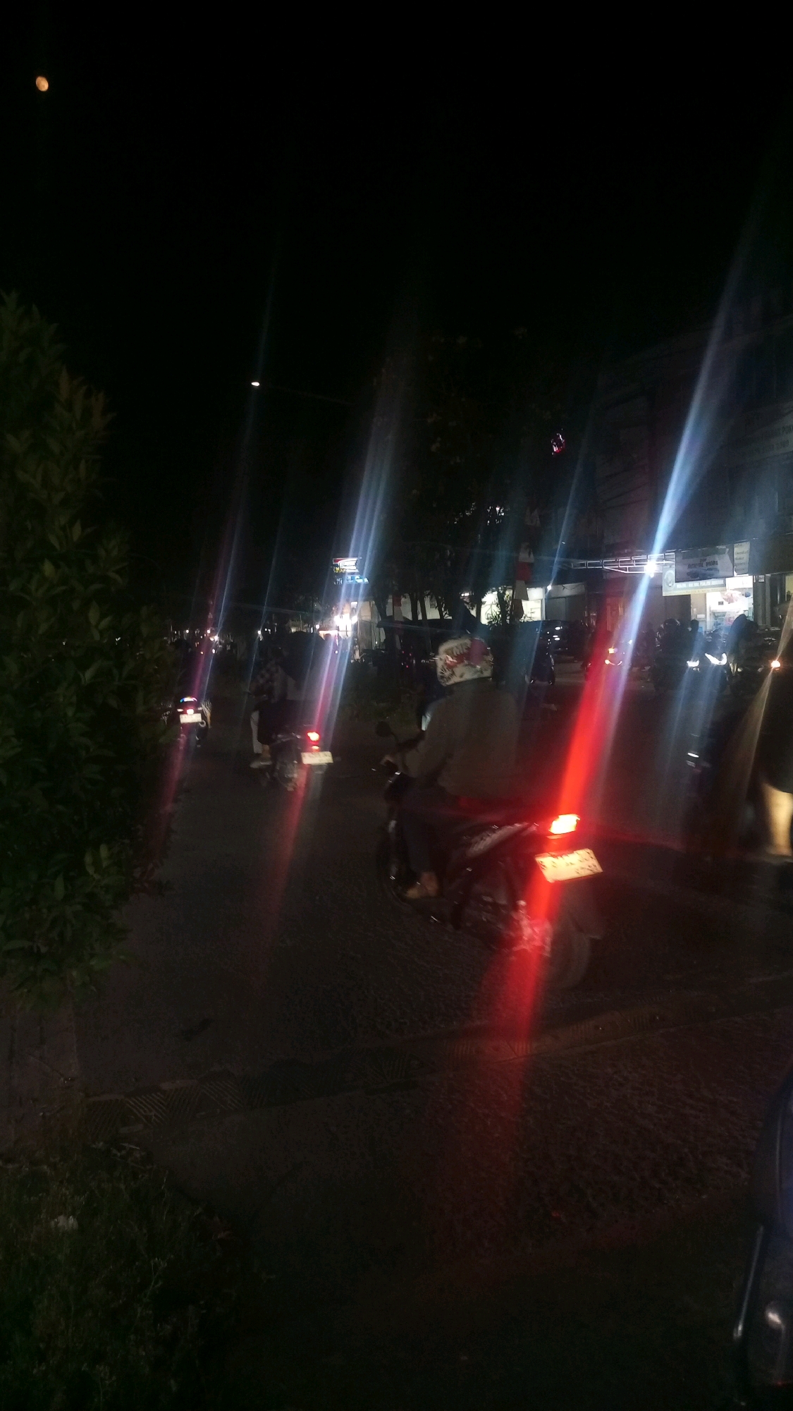 The image captures a night scene in Babakan Asem, Indonesia, likely on a street with moderate pedestrian and vehicle traffic. The foreground is dominated by a dark, asphalt road, marked with what appear to be dashed white lines and possibly some subtle texture variations. To the left, a dense, dark green bush or small tree is visible, with a few small, white debris pieces on the ground beneath it.
The central focus of the image is a person riding a motorcycle, viewed from the rear. The rider wears a light-colored jacket and a helmet with a patterned design. The motorcycle itself has a red taillight illuminated, casting a bright red streak of light across the road. A license plate, partially obscured, is visible on the rear of the motorcycle.
Further down the road, several other motorcycles are present, some with their taillights on, creating multiple red light streaks. These motorcycles are either parked or in motion, with figures of riders visible on some.
In the background, buildings line the street, with illuminated signs and windows suggesting commercial establishments or residences. The lighting in the scene is characterized by strong light streaks, likely from streetlights and vehicle headlights, creating lens flare effects. These lights illuminate parts of the road, buildings, and foliage. The overall atmosphere is that of a busy street at night.