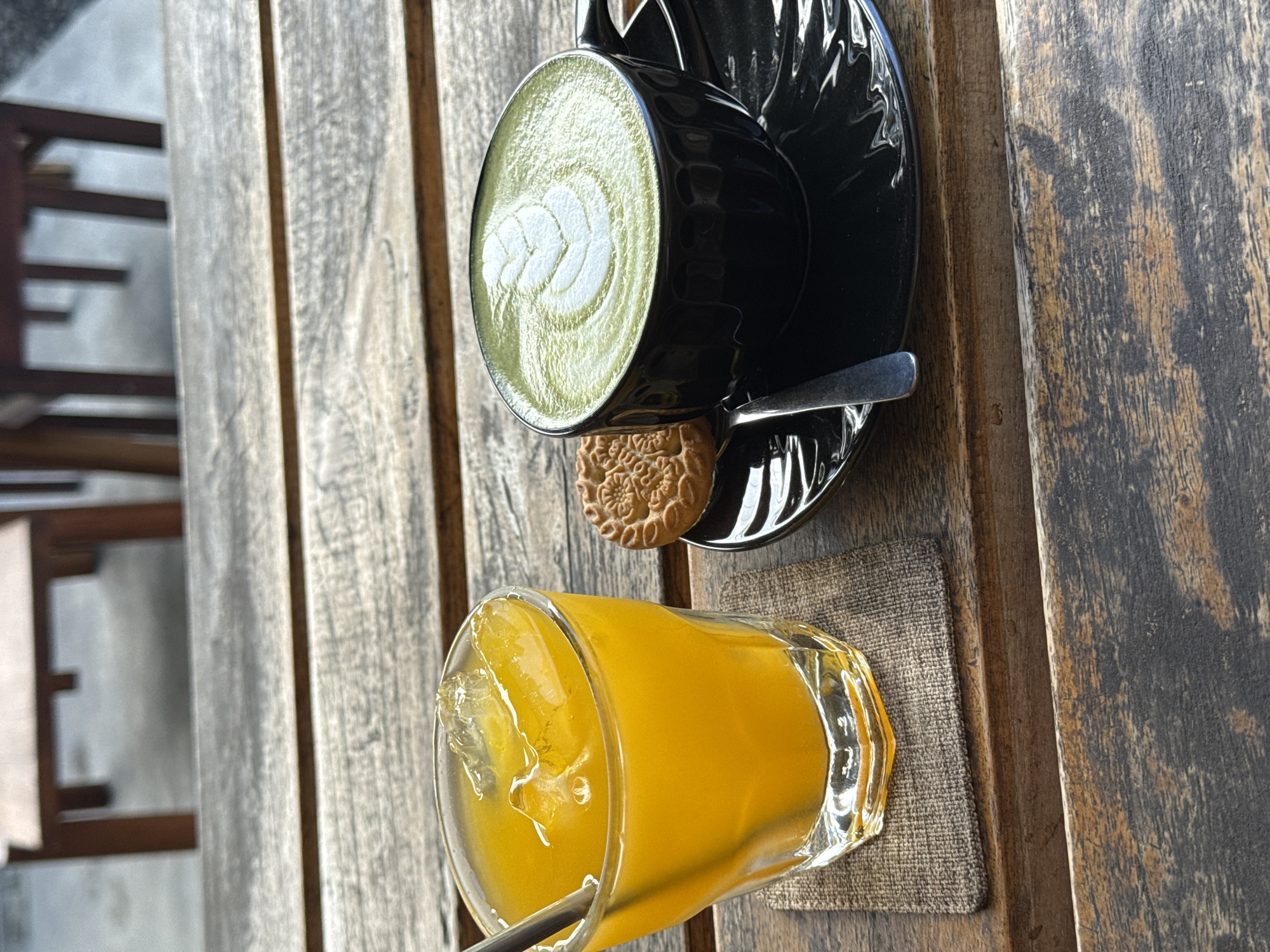 A top-down shot presents a wooden plank surface. On the upper right, a black cup filled with a green matcha latte sits on a matching saucer. Beside it, a small round biscuit rests on the saucer. To the lower right, a glass of orange juice with ice sits on a gray woven coaster. A metal straw is visible in the glass. A portion of a wooden chair can be seen in the upper left. The location appears to be in the city of Unknown, Unknown.