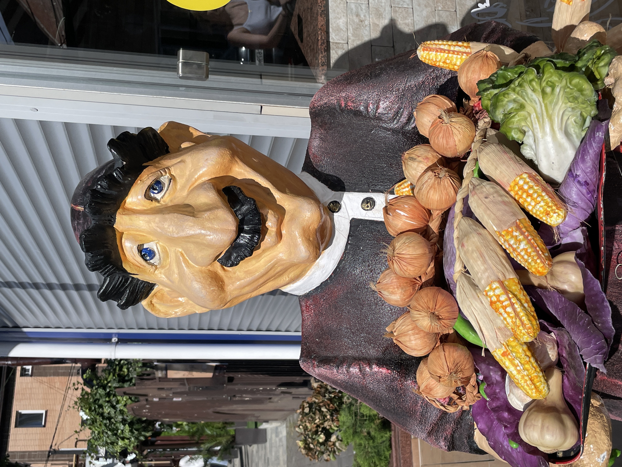 This image shows a statue of a man holding a tray of vegetables. The statue is life-sized and made of a brightly colored material, possibly plaster or fiberglass. The man has a large head with exaggerated features, including a prominent nose, thick black mustache, and wide blue eyes. He is wearing a dark red jacket with a white collar and black buttons.
The tray he holds contains a variety of produce, including several ears of corn, heads of garlic, onions, red cabbage, and a head of lettuce. The vegetables appear to be made of artificial materials as well, likely to mimic real produce.
In the background, there is a building with light-colored siding and a window. To the left of the statue, there is a fence and some greenery. The scene appears to be outdoors, possibly on a street or in front of a shop.
While the image does not provide explicit clues to its location within Sochi, Russia, such statues are often found in public spaces, markets, or as decorative elements outside establishments like restaurants or cafes. The style of the statue and the presentation of produce are consistent with decorative elements found in various tourist locations.