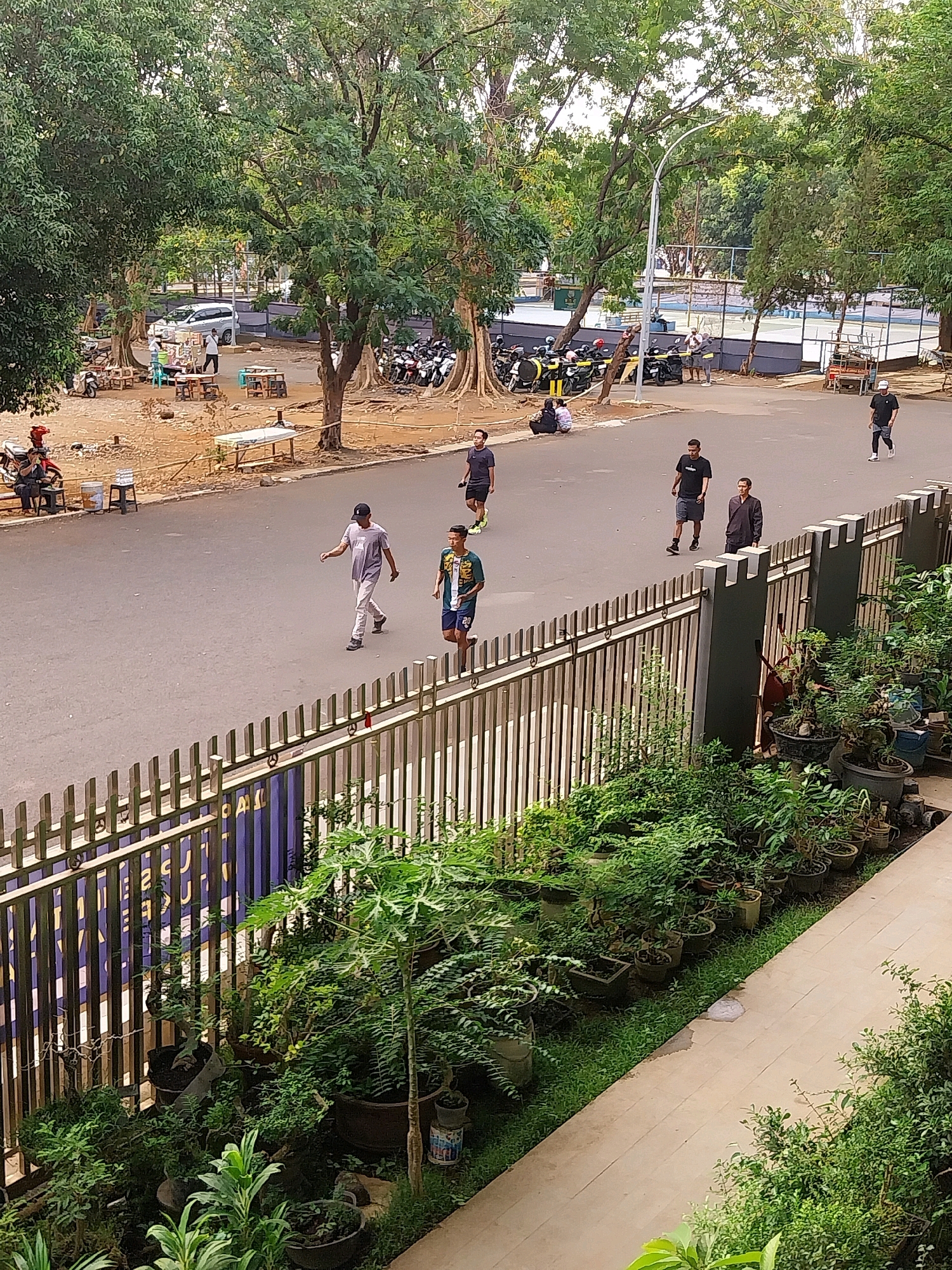 The image captures a street scene in Cirebon, Indonesia, featuring a paved road, a metal fence, and a variety of trees and vegetation. On the road, several individuals are present, some walking and others seemingly engaged in casual activity. In the background, there are parked motorcycles, a tennis court, and a few structures that might be food stalls or small shops. The overall environment suggests a public outdoor area, possibly a park or a recreational space within the city. The time of day appears to be late afternoon, given the lighting conditions.