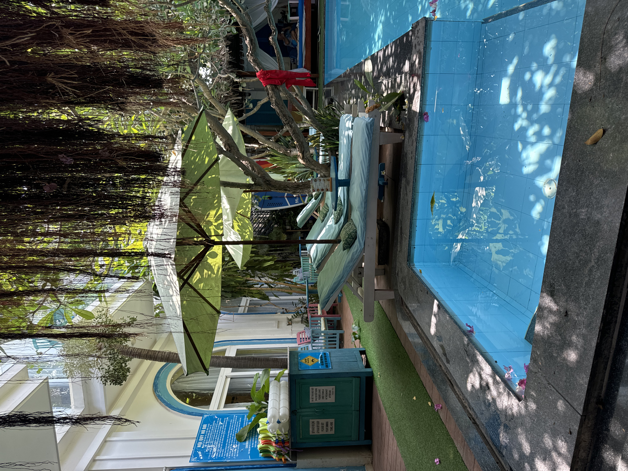A resort in Mũi Né, Vietnam, features a swimming pool with blue tiles. Adjacent to the pool are lounge chairs shaded by a green umbrella. A small blue cabinet stands nearby, next to a sign with swimming pool rules.