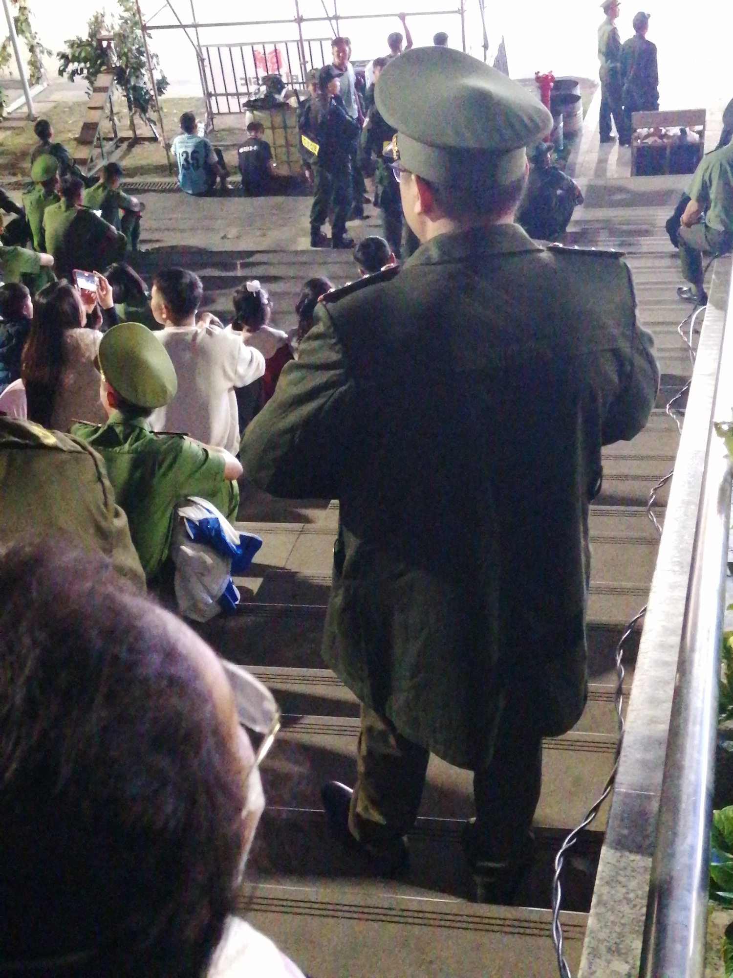 The image depicts a group of people, some in military-style uniforms, gathered on outdoor stairs at dusk. The setting appears to be a public area, possibly a park or a stadium, with a metal railing and a partially visible fence in the background. Some individuals are seated on the steps, while others are standing. A prominent figure in the foreground wears a dark green uniform and a matching cap, facing away from the camera. The overall mood is subdued, with artificial lighting casting shadows on the scene. There are no identifiable landmarks that specifically place the scene in Da Lat, Vietnam; however, the presence of uniformed individuals and the outdoor setting are consistent with various public gatherings that might occur in the city.