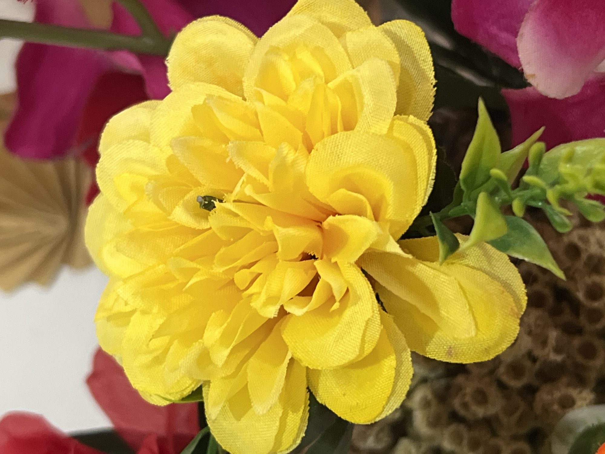 The image shows a close-up of a bright yellow artificial flower with many layered petals. A tiny dark insect is visible in the center of the flower. The flower is part of a bouquet, with blurry red and magenta flowers visible in the background, along with some greenery. The background also features a blurry, tan, fan-like object and a textured brown area that could be part of another artificial plant. The lighting is bright, illuminating the texture of the fabric petals. The setting appears to be indoors, possibly a home or a shop display, with no discernible elements to indicate a specific location in Denpasar, Indonesia.