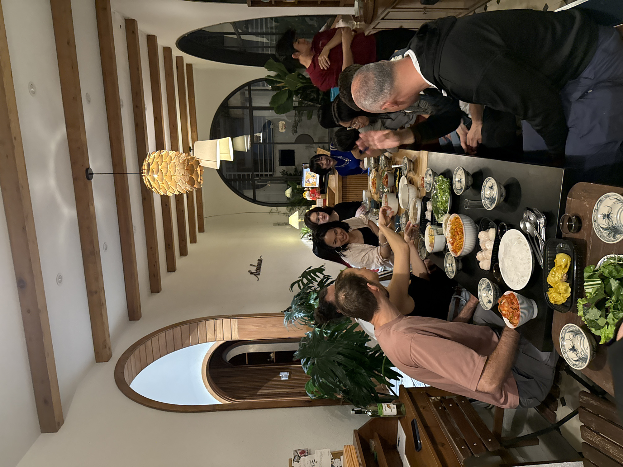 The image shows a group of people, approximately ten individuals, gathered around a dark-colored dining table laden with various dishes and bowls of food. The setting appears to be a dining room with warm, ambient lighting, featuring wooden beams on the ceiling and arched doorways. Several large potted plants are visible, contributing to a cozy atmosphere.
The individuals are diverse in age and gender, dressed in casual attire. They are engaged in eating and conversing, with some individuals looking at each other and interacting. One person in a pink shirt is seen with their back to the camera, while others are partially visible or in profile. The food on the table includes what appears to be rice, vegetables, and several bowls of what might be kimchi or other pickled vegetables, as well as some dumplings.
The architectural style, with its arched openings and wooden elements, suggests a modern home or restaurant. The presence of a bottle of white wine on a nearby wooden desk, along with some papers, indicates a social gathering. The overall impression is one of a communal meal and pleasant social interaction.
While the image does not explicitly state the location within Lam Vien - Da Lat, Vietnam, the style of the architecture and the presence of lush greenery could be consistent with properties found in such a city known for its cool climate and natural beauty. The atmosphere is relaxed and convivial.