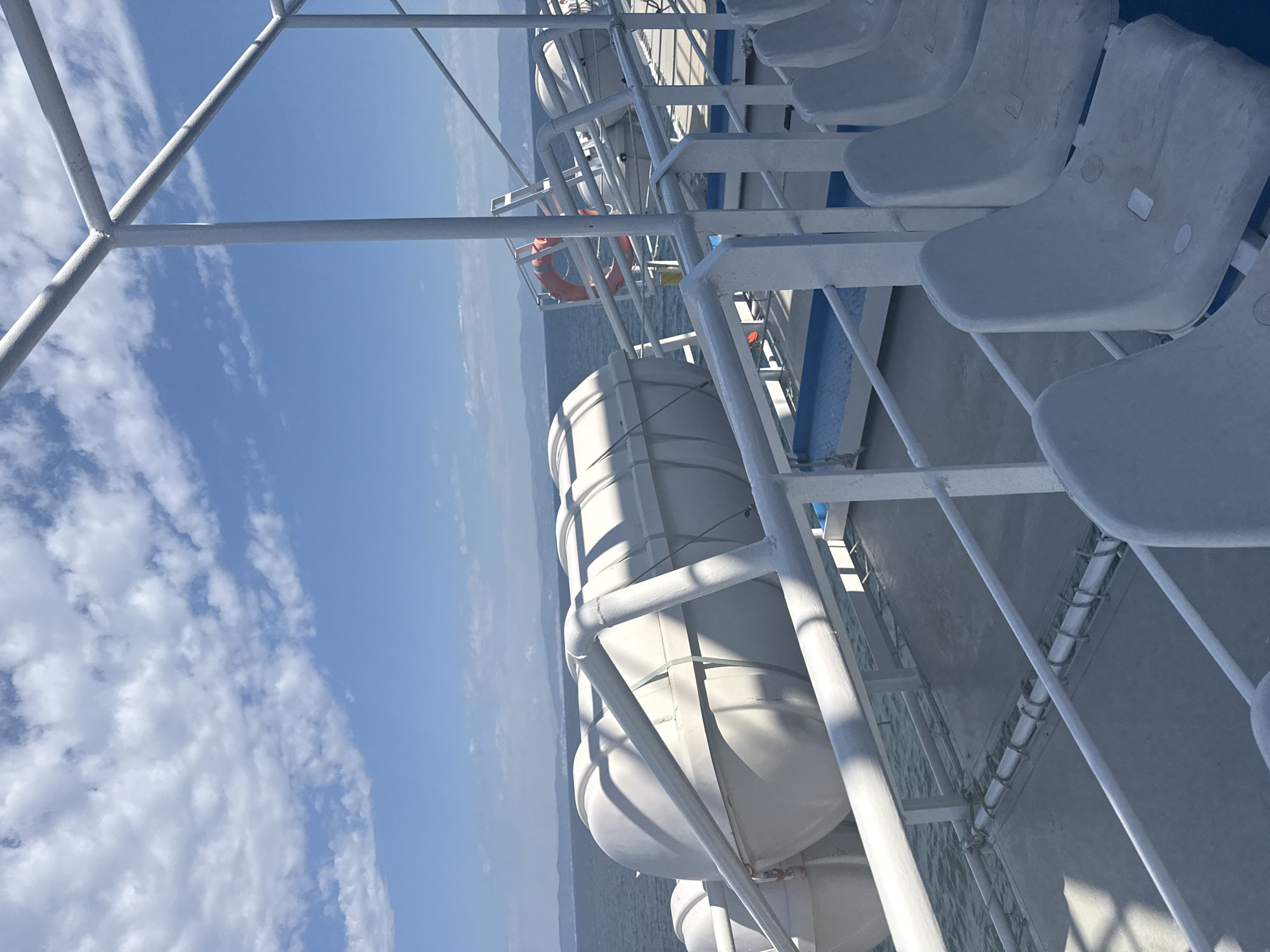 The image depicts the deck of a ferry with rows of empty white seats in the foreground. In the middle ground, several large white, cylindrical life rafts are secured to the deck, with a bright orange life preserver visible further down the railing. The background shows a calm sea under a blue sky with scattered white clouds. Distant, hazy landmasses can be seen on the horizon. The scene appears to be a sunny day at sea. This setting is consistent with the ferry terminal in Prinos, Greece, a common departure point for sea travel.
