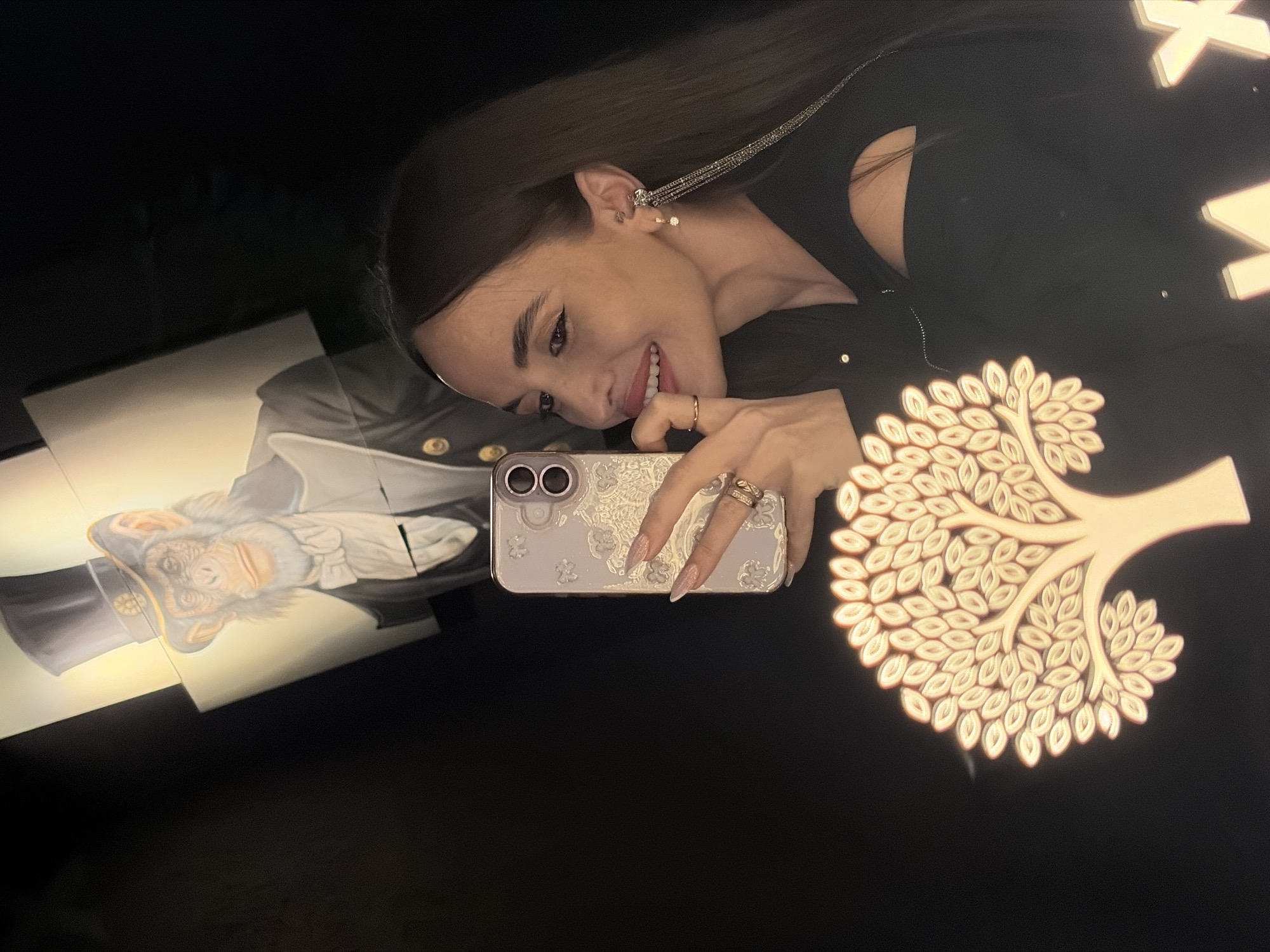 The image shows a young woman taking a selfie in front of an art installation. The woman has long, dark hair and is wearing a black top. She is holding a smartphone with a patterned case. The art installation includes a portrait of an ape wearing a formal military uniform and a stylized tree made of light-colored, possibly illuminated, elements. The setting appears to be indoors, with dark walls. There are also illuminated letters partially visible in the background, which may indicate a sign or logo.
The location is the Saint Petersburg Museum of Emotions in Saint Petersburg, Russia.