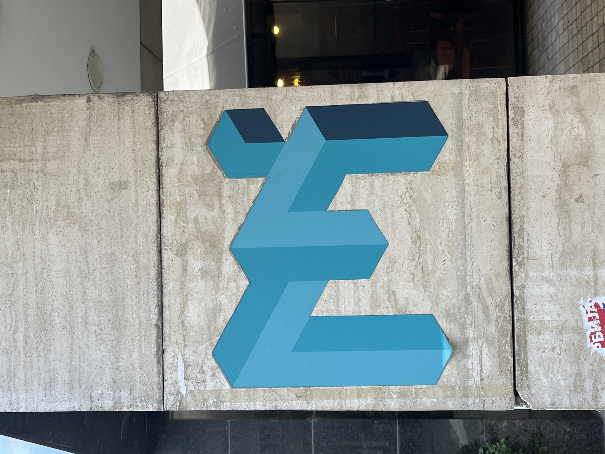 Here is a description of the image:
A blue, geometric design resembling a stylized "E" or "Sigma" is painted on a concrete structure. The design incorporates a three-dimensional illusion using shades of blue to suggest depth and perspective. The concrete structure appears to be a part of a building facade or architectural detail, given the presence of dark material. The location includes a sticker to the right of the design with the letters "BAJA." This sticker, along with the design and architectural style, suggest that the scene is likely located in the city of Belgrade, Serbia.
