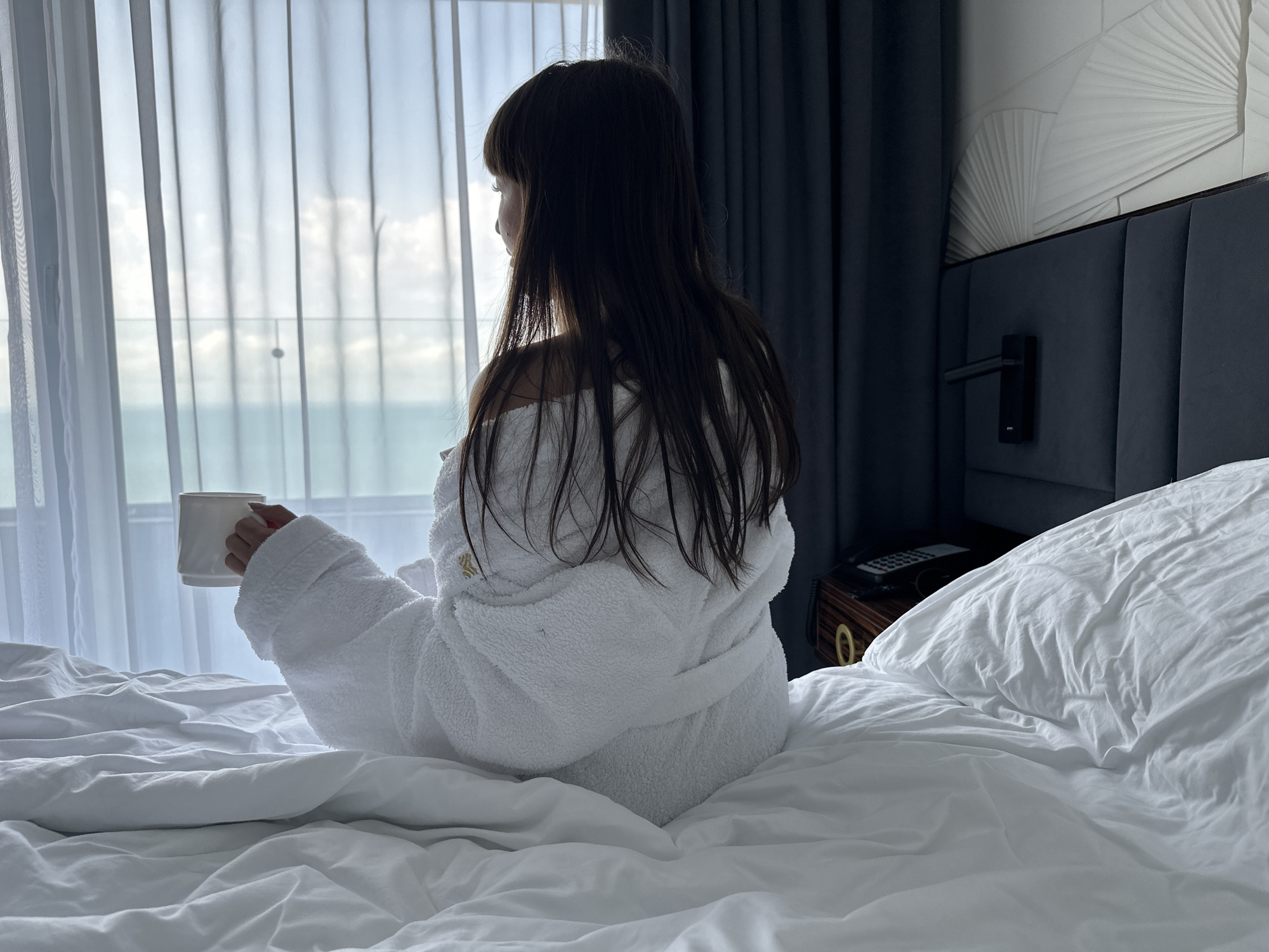 This media file depicts a serene scene inside what appears to be a hotel bedroom in Sochi, Russia. The primary subject is a person, seen from behind, with long, dark hair, sitting up in a large bed covered with white bedding. The person is wearing a fluffy, white bathrobe and holds a white mug in their left hand, gazing out a window.
The window is covered by sheer white curtains, through which a bright sky with scattered clouds and a wide expanse of the sea or ocean are visible. The natural light from the window brightly illuminates the scene, creating a sense of calm.
To the right of the bed, part of a dark, upholstered headboard is visible, extending up to a decorative wall panel featuring a white, fan-like, textured design. A small, dark-colored nightstand is partially visible, holding a remote control and what might be a telephone. The bed itself is draped in white sheets and duvet, showing natural wrinkles.
The activity taking place is the person peacefully observing the outdoor view, possibly enjoying a morning beverage. The overall atmosphere suggests relaxation and contemplation, likely during the daytime given the bright light from outside. No discernible text is visible on any of the objects or surfaces.