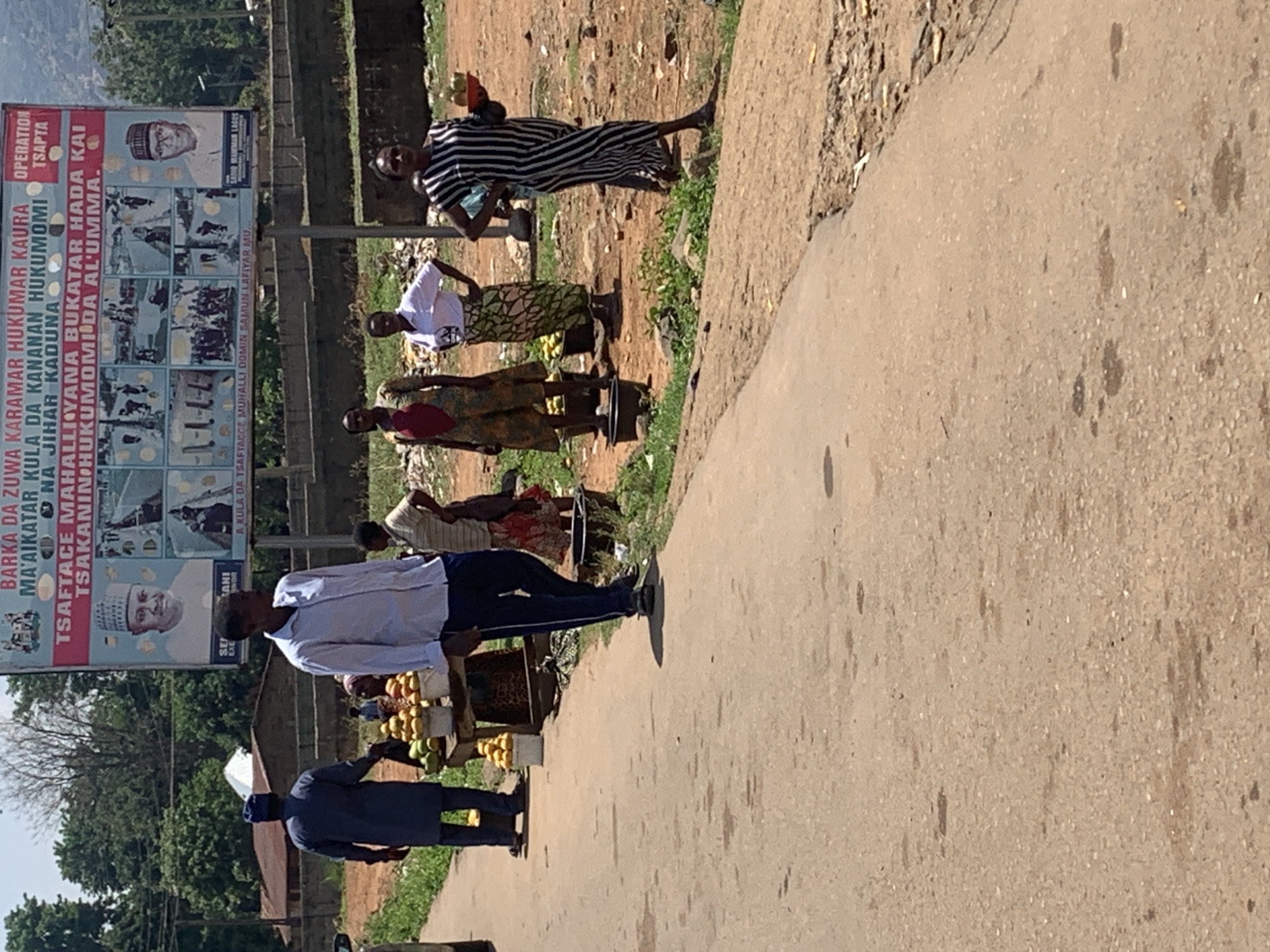 The image depicts a street scene in Kukum, Nigeria. A large political billboard stands beside the road featuring pictures of people and Hausa script. Several people stand beside the road selling various wares, including fruit. The road is paved and the surrounding environment is dirt and vegetation.