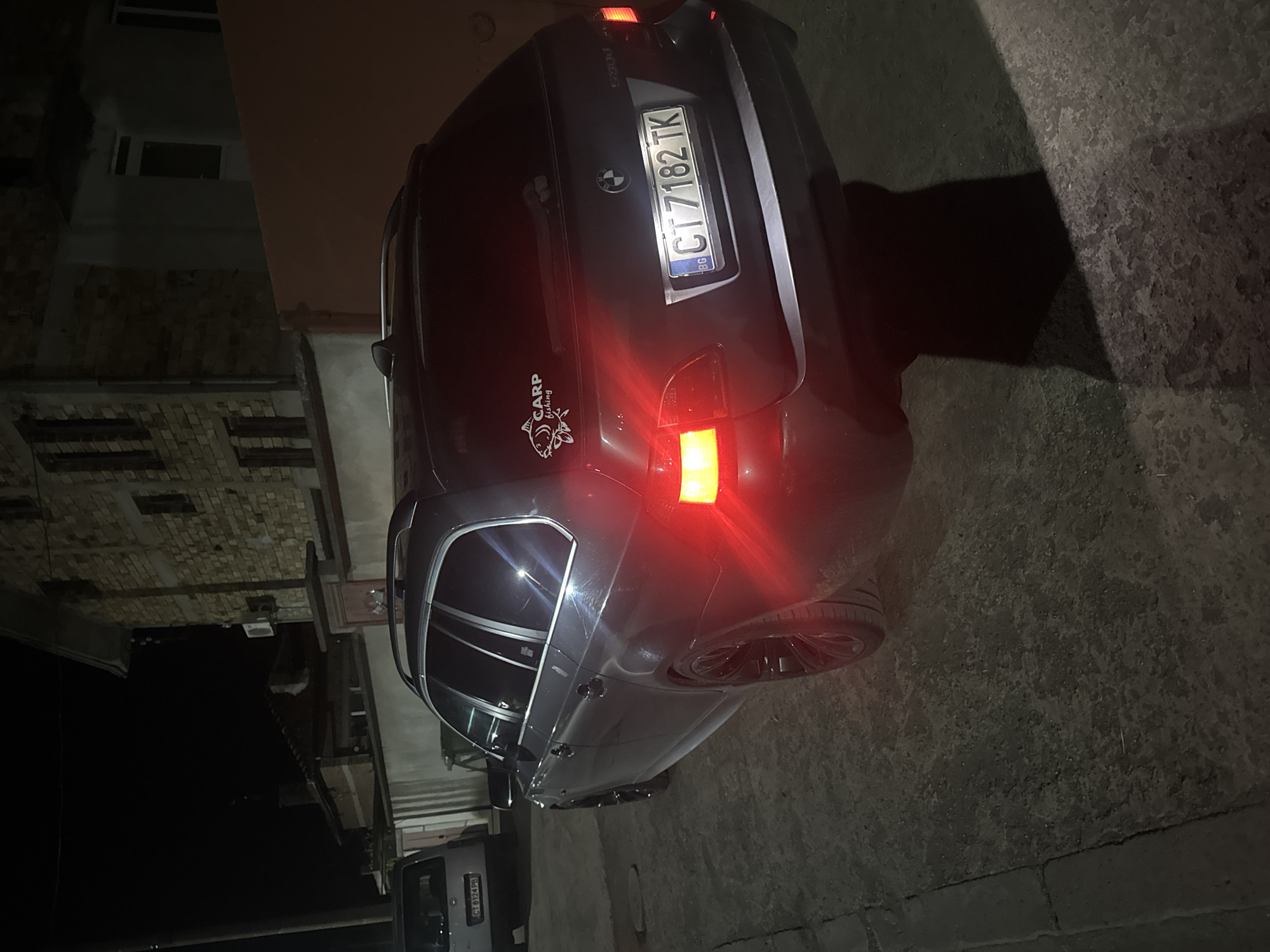 At night in Sarnevo, Bulgaria, a dark gray BMW station wagon with license plate CT 7182 TK is parked on a paved surface. A "CARP fishing" sticker is on the rear. Part of a multi-story building with patterned brickwork is visible in the background. Another car with license plate CT 6174 PB is parked behind the BMW. The BMW's right taillight is illuminated.